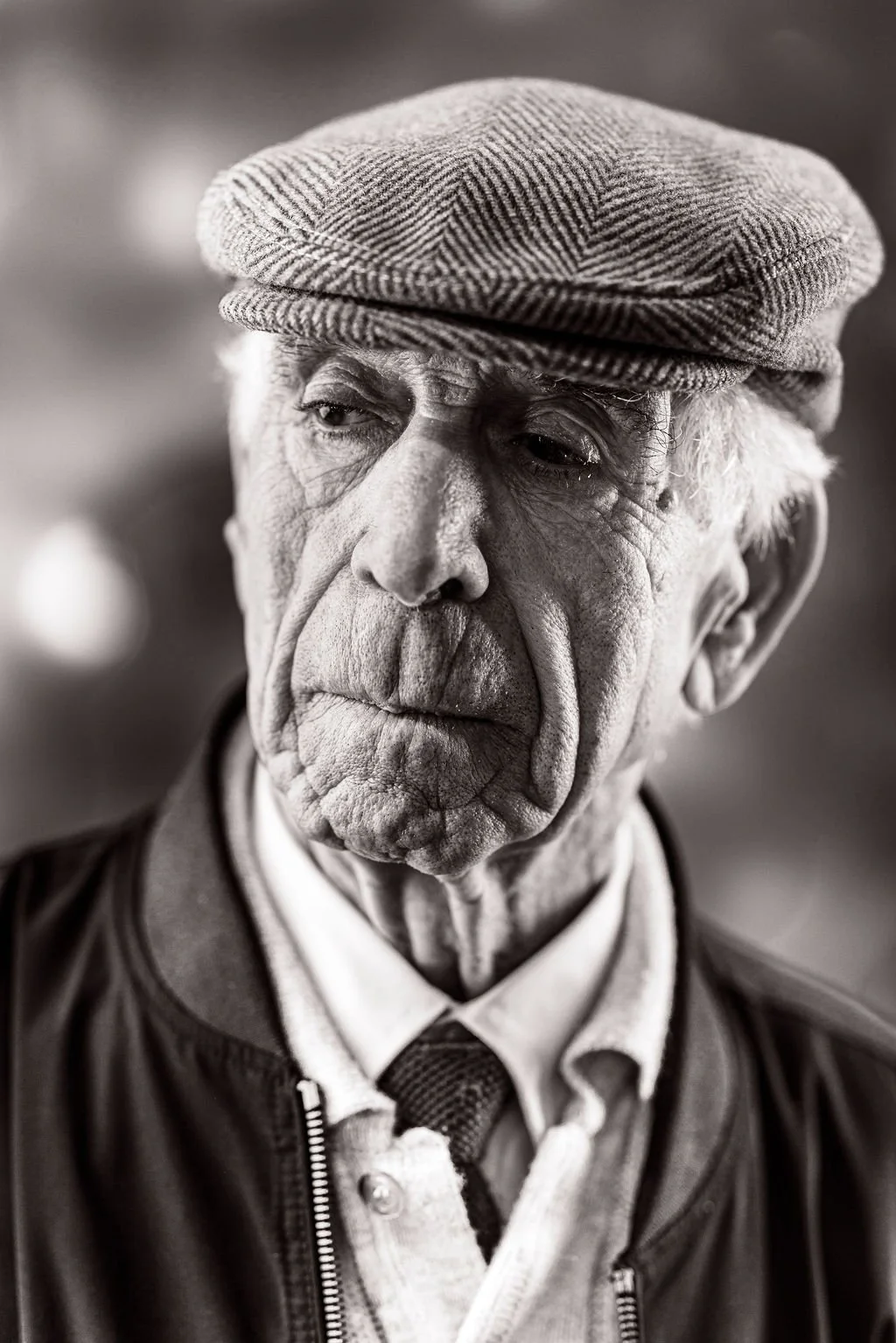 Close-up black and white photo of an elderly man with wrinkles, wearing a flat cap and a jacket, looking contemplative.