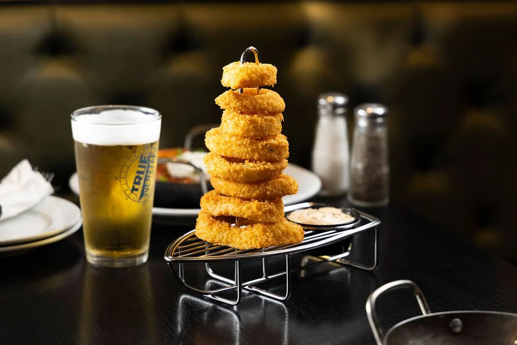 A tower of crispy fried onion rings on a wire rack, with a small container of dipping sauce, a glass of beer, and condiments on a dark table in a restaurant.