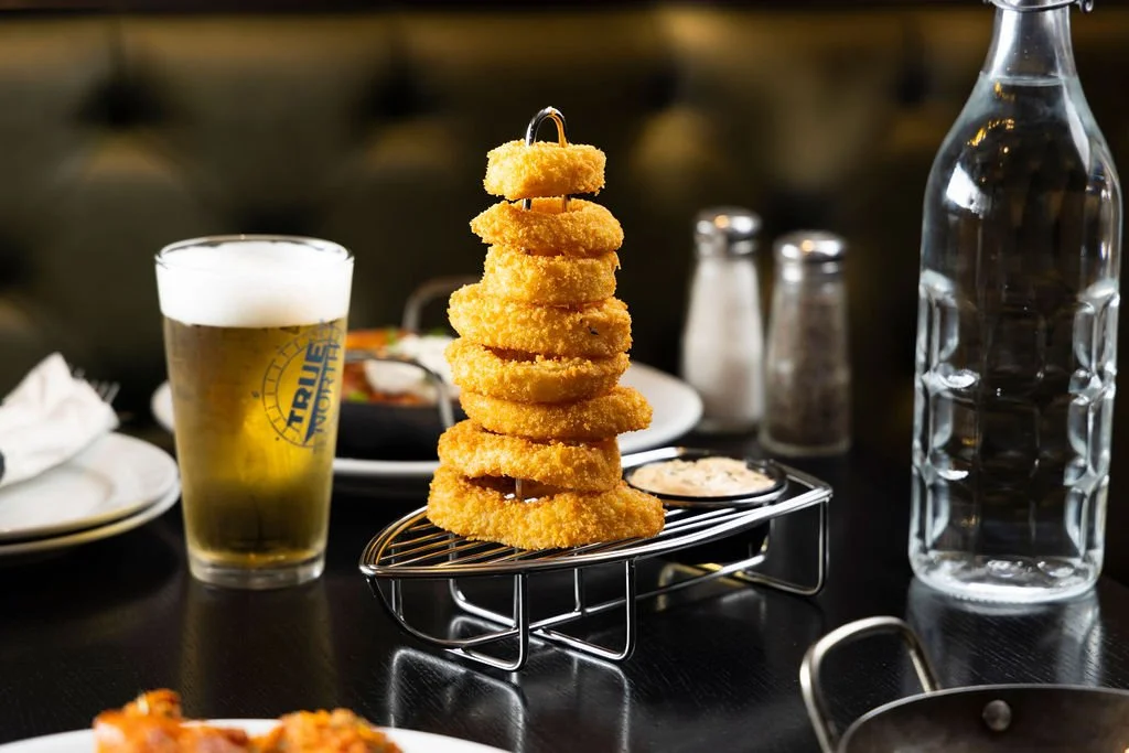 A tower of fried onion rings on a serving rack, with a glass of beer, a water bottle, and condiments on a dark table at a restaurant.