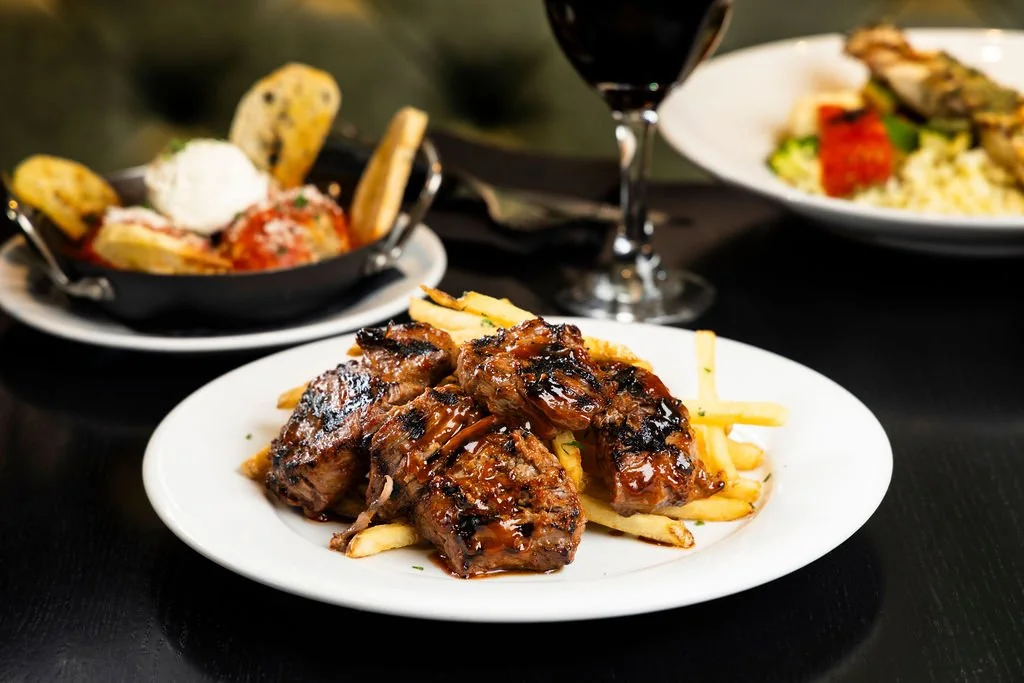 Plate of grilled ribs served with French fries on a white plate, with a side dish of baked spaghetti, a glass of red wine, and another plate with salad and what appears to be baked fish in the background.