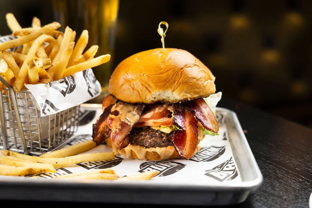 A gourmet burger with bacon, tomato, lettuce, and cheese on a bun, served with fries on side on a metal tray.