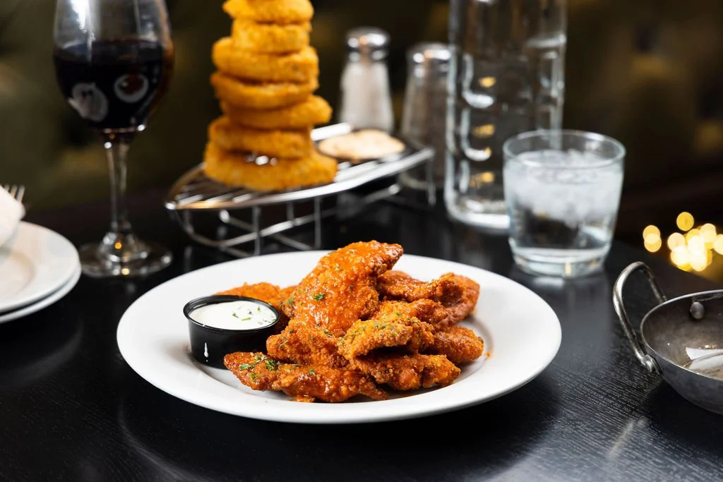 Plate of fried chicken tenders with dipping sauce, a stack of onion rings in a wire basket, a glass of red wine, a glass of water, and a glass pitcher on a dark table.