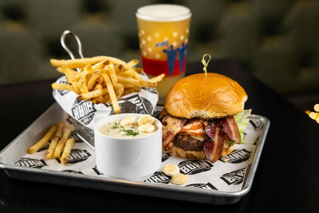 Tray with cheeseburger topped with bacon, French fries, a cup of chowder with clams, and a craft beer in a colorful glass