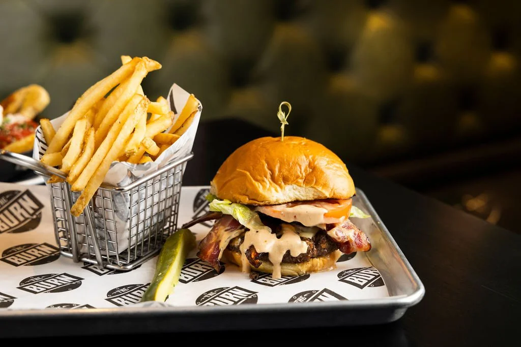 A burger with lettuce, tomato, cheese, a beef patty, bacon, and sauce in a bun, served with a side of French fries and a pickle spear on a tray.