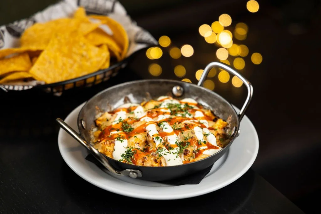 A small black skillet containing loaded nachos topped with cheese, sour cream, chopped herbs, and hot sauce, placed on a white plate. In the background, there is a basket of tortilla chips with a black and white checkered paper liner. The scene is il