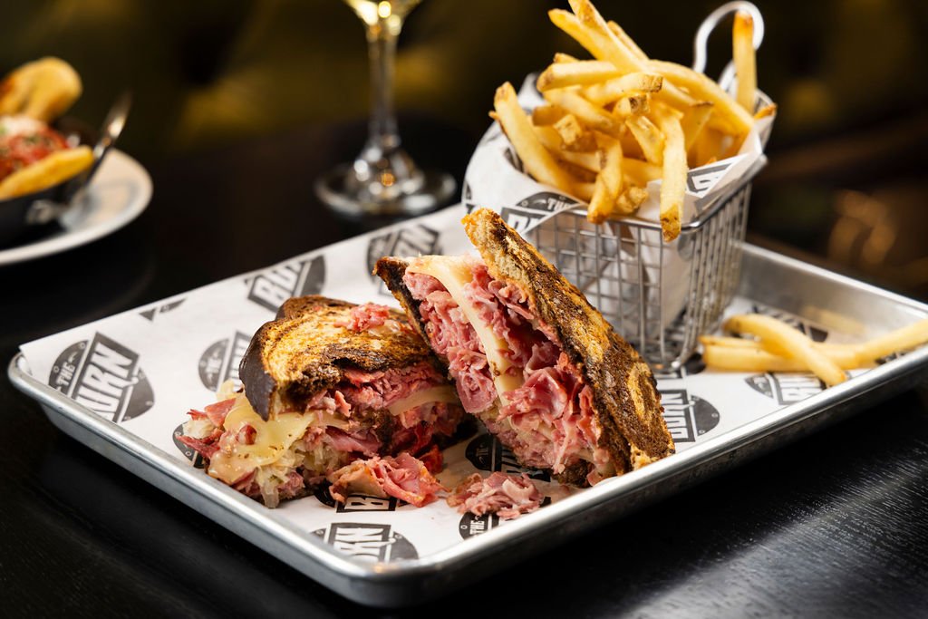 Half of a grilled Reuben sandwich with melted cheese and corned beef, served with a side of French fries in a small wire basket on a tray with paper liner, in a restaurant setting.