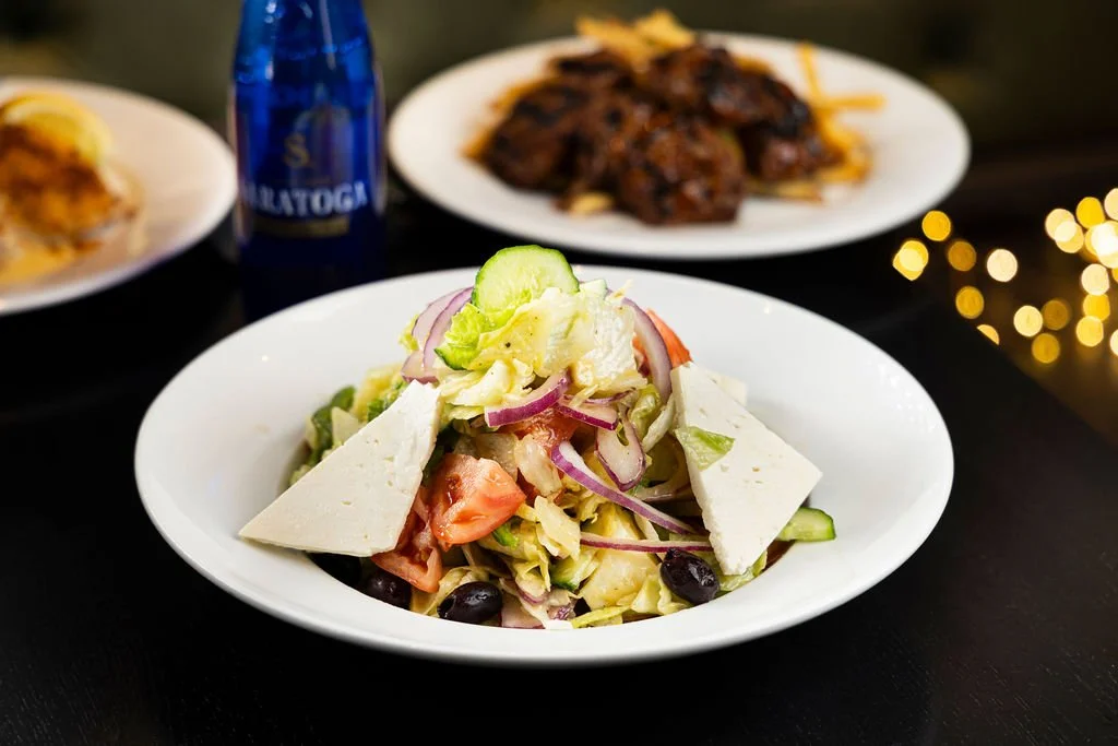 A plate of salad with lettuce, cucumber, tomato, red onion, black olives, and slices of feta cheese in the foreground. In the background, there are plates with meat dishes and a bottle of Saratoga bottled water.