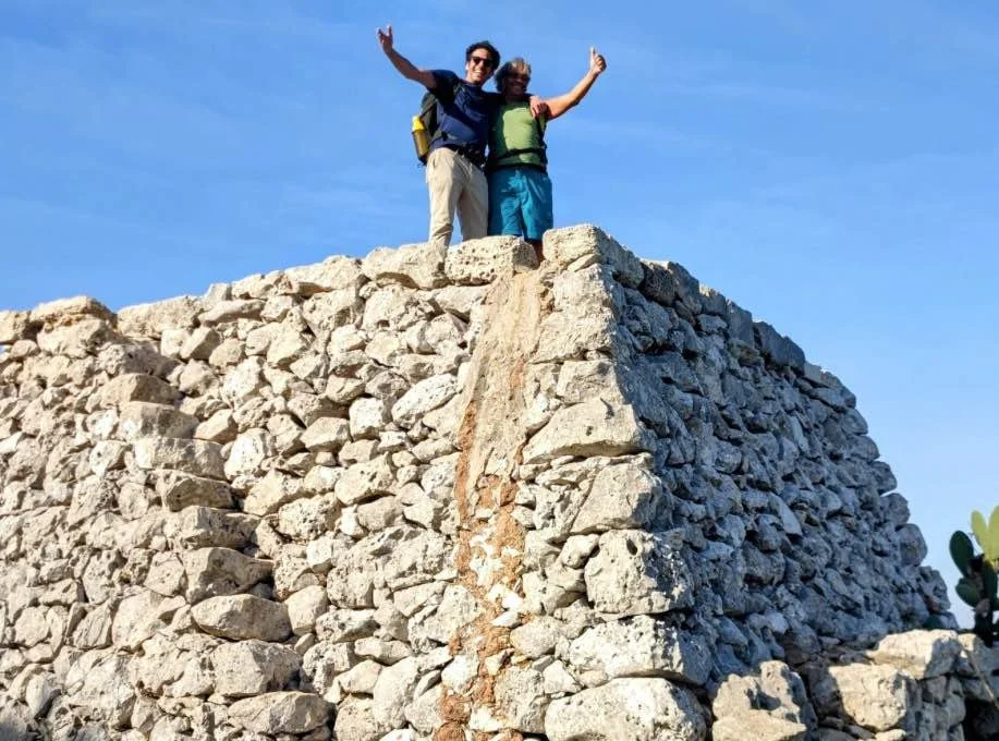 Due escursionisti con le mani alzate sopra una pagliara lungo il Cammino del Salento.