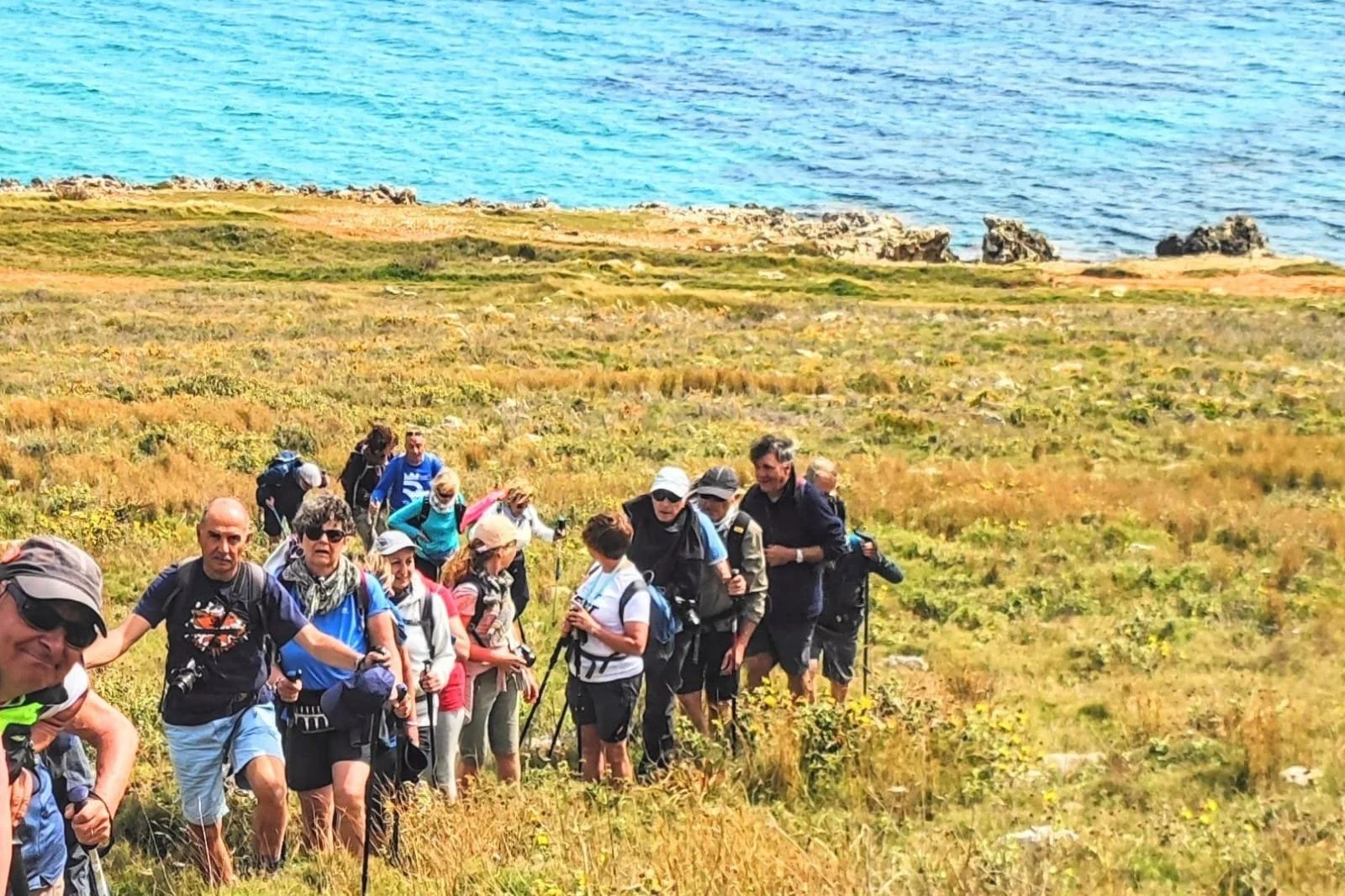 Gruppo di camminatori in fila su un prato vista mare lungo il Cammino del Salento