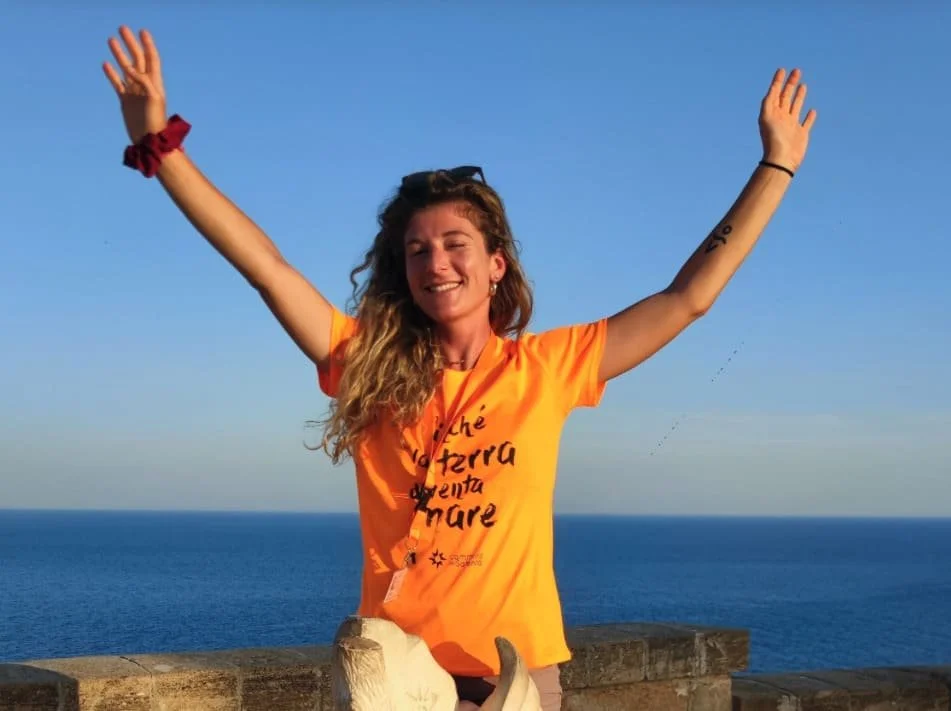 Ragazza con la maglia arancione del Cammino del Salento, mani alzate e sorriso davanti al mare.