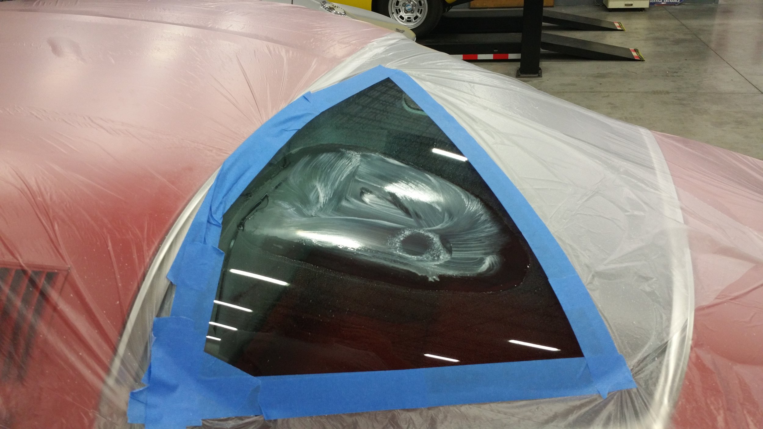 Car window masked with painter's tape and plastic, partially cleaned with a cream or polish.