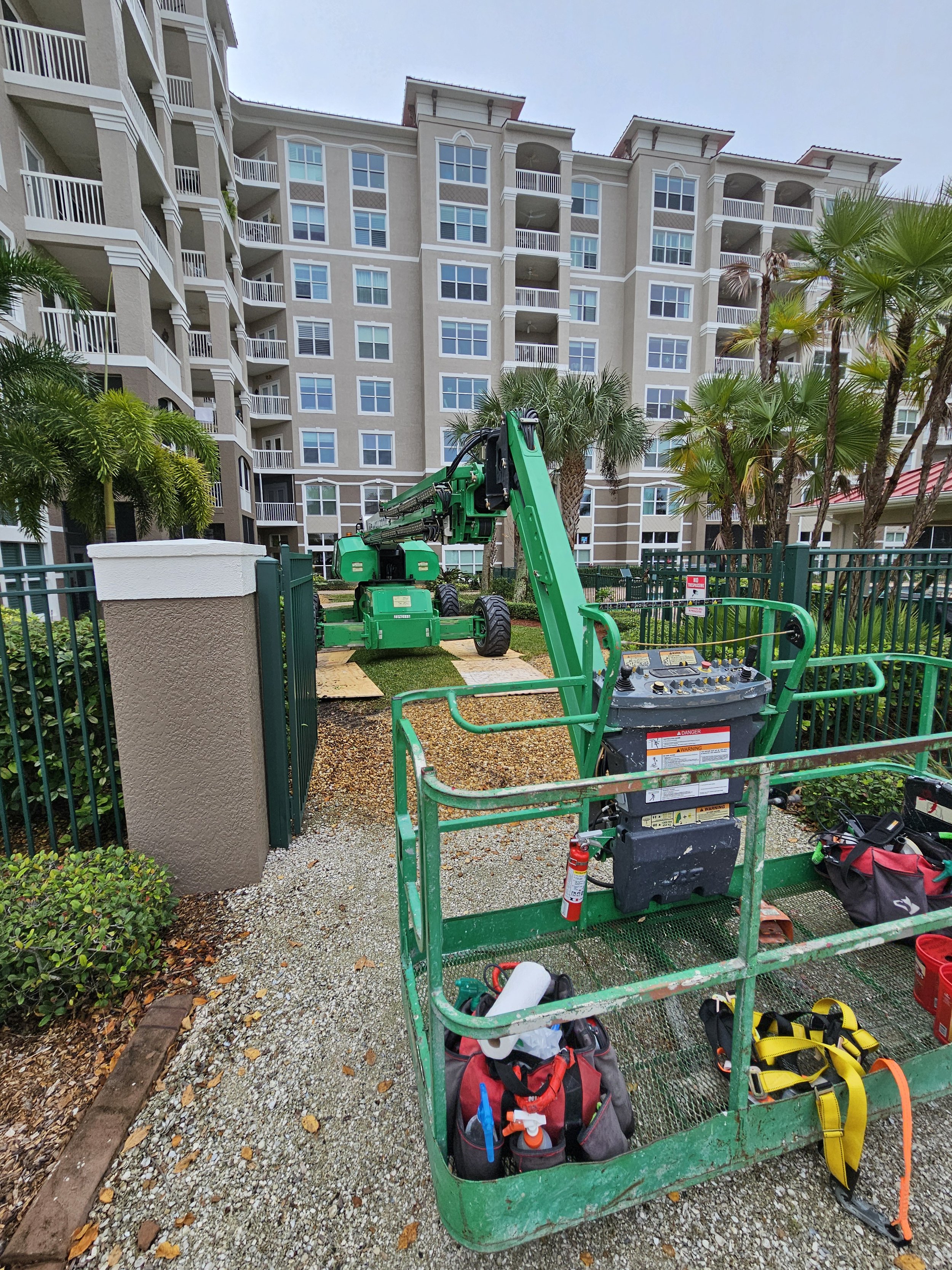 Construction site with green boom lift in front of a multi-story residential building, surrounded by palm trees and landscaping, with tools and equipment visible in the lift's basket.
