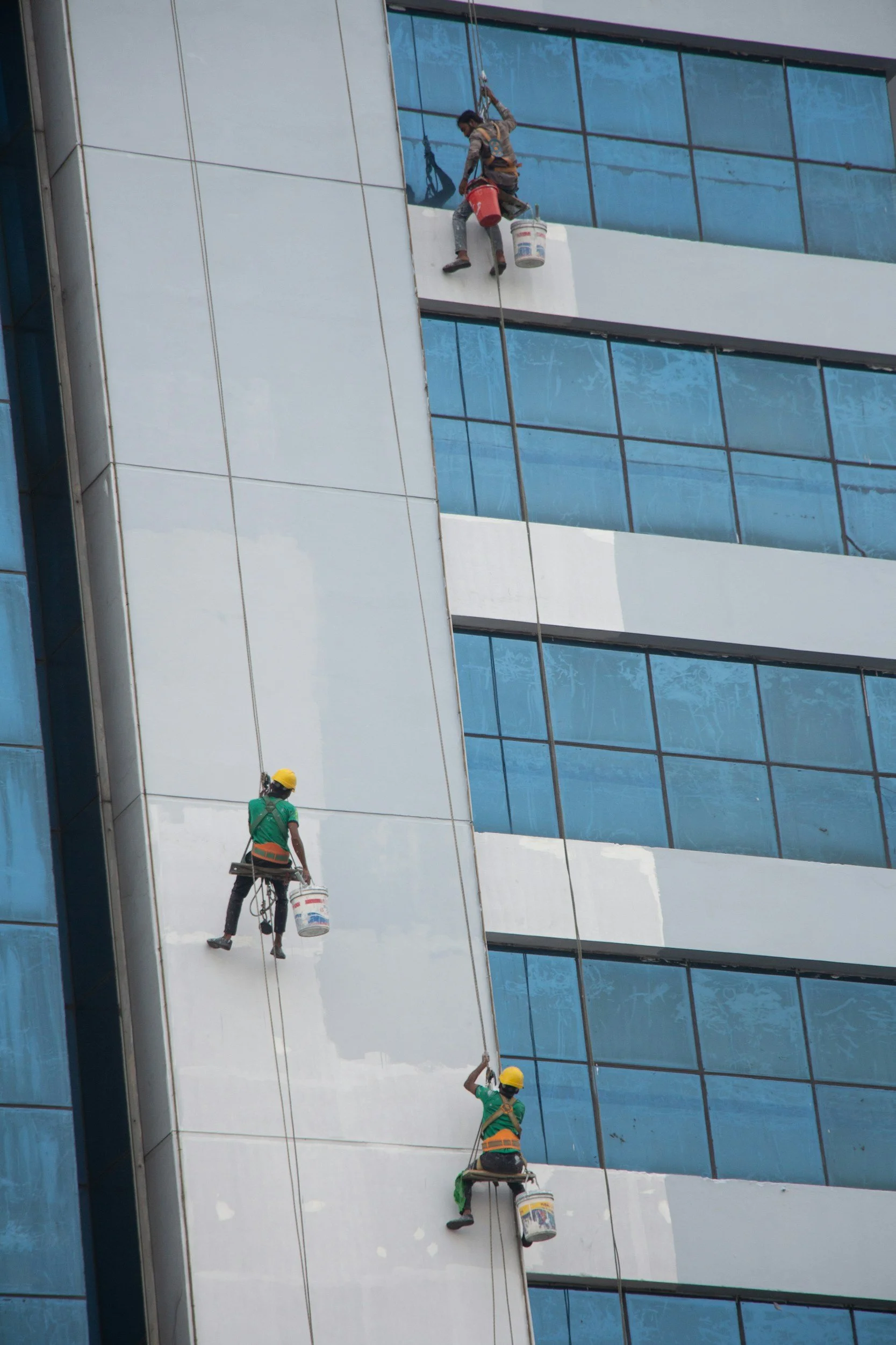 Painters on a bosun's chair working alongside glass and cladding on a commercial high-rise, the same job site conditions where Glass Restoration Inc. is often called in for accidental glass damage