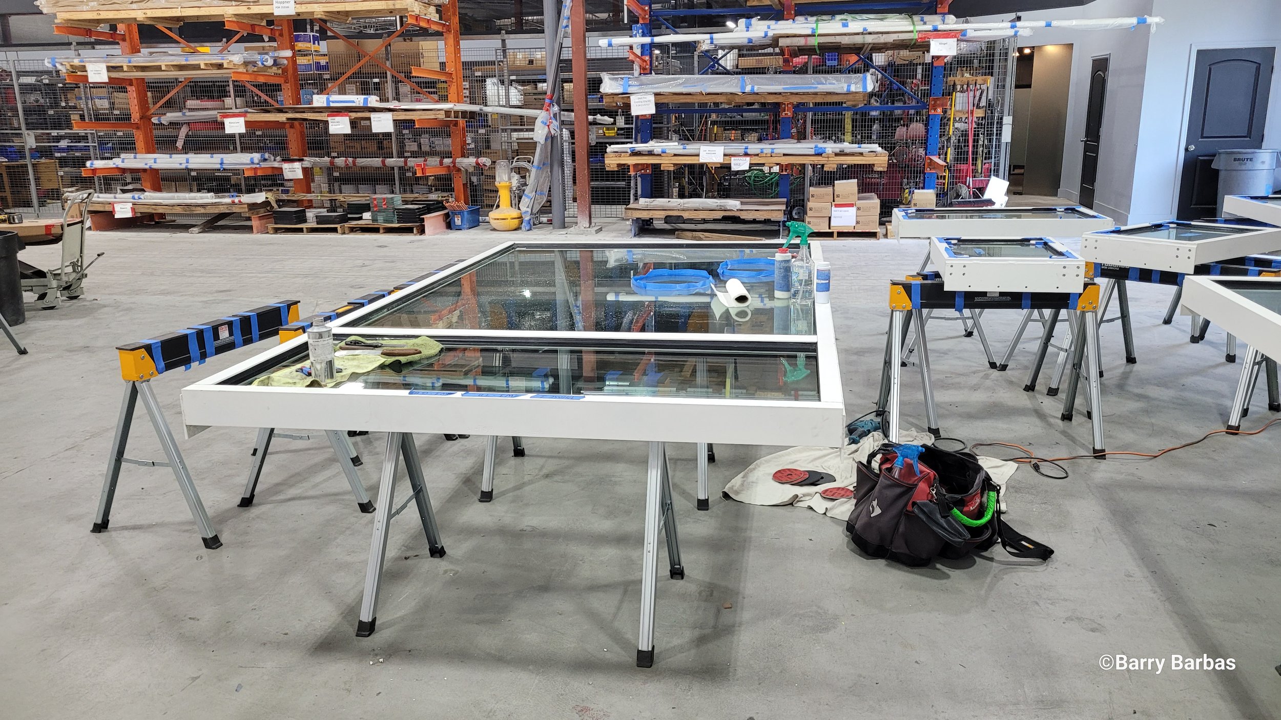 Workshop with large glass panel on sawhorses, shelves with materials, and tools scattered on the floor.
