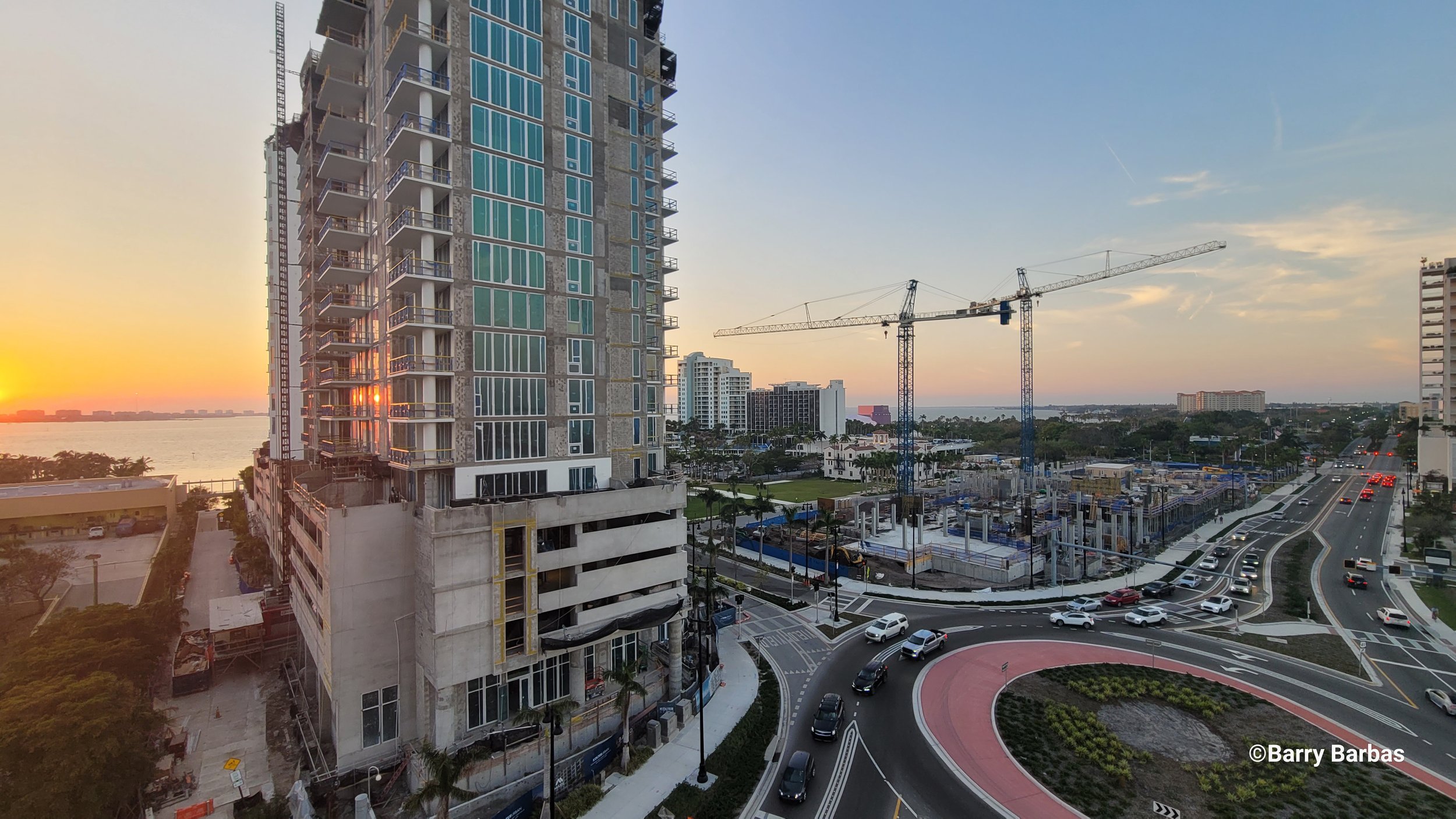 Construction site with a high-rise building, cranes, and a roundabout, overlooking a waterfront at sunset.
