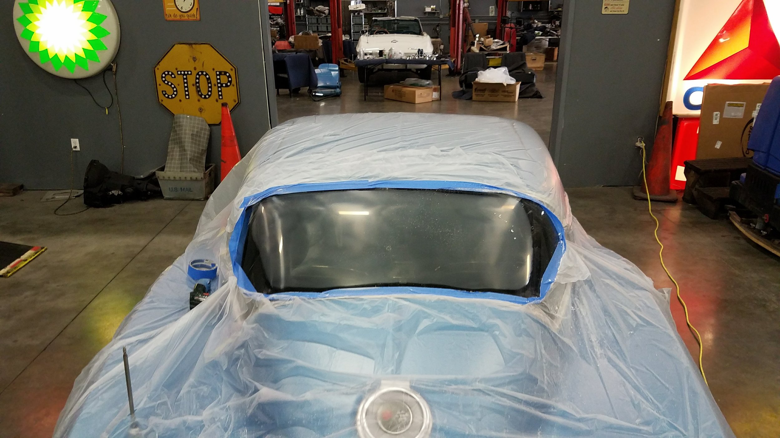 Car covered with plastic for painting in garage, surrounded by automotive signs and workshop equipment.