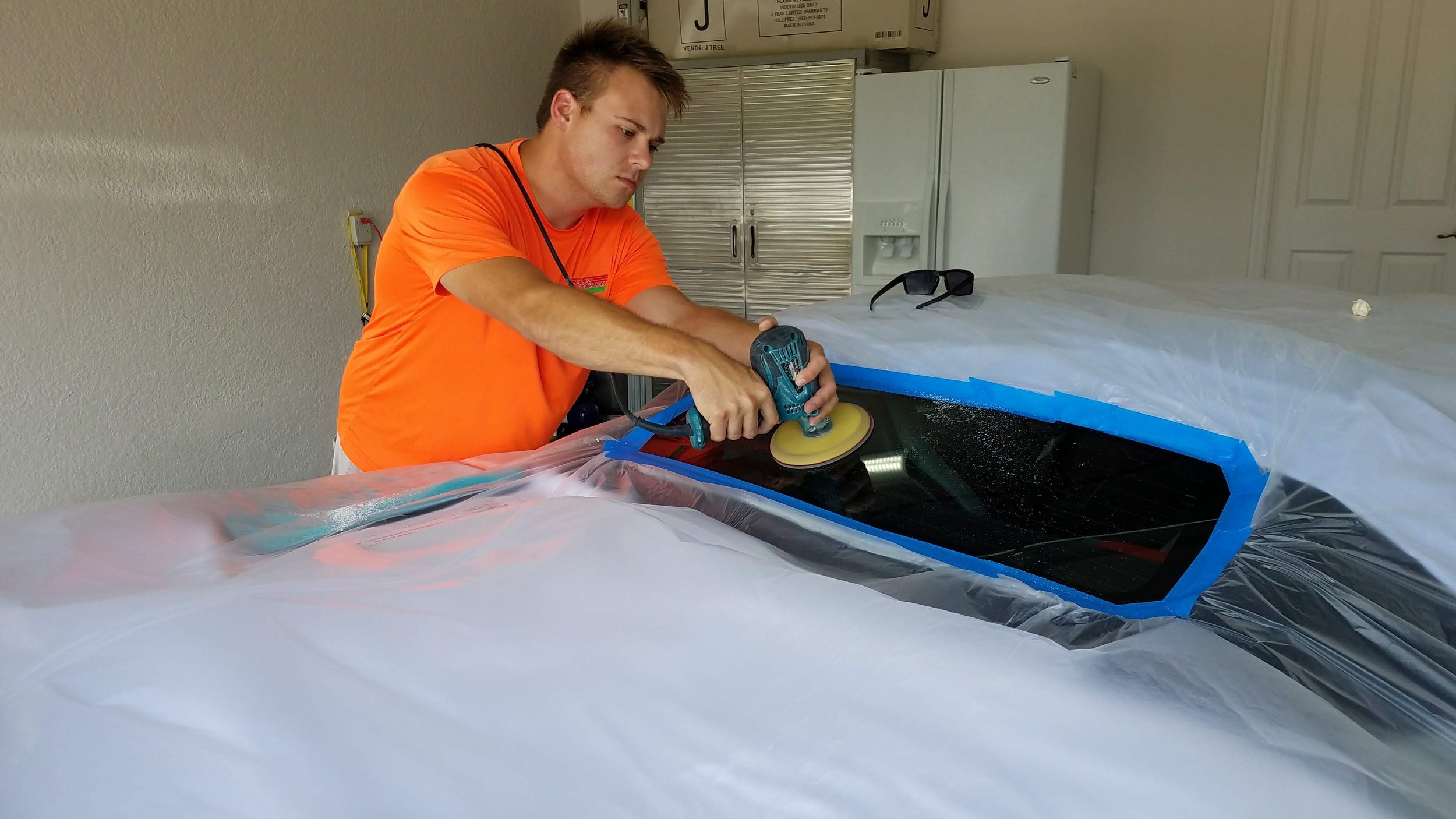 A person wearing an orange shirt uses a power tool to polish a vehicle's rear window, which is taped off with blue tape, while the rest of the car is covered in a protective sheet.
