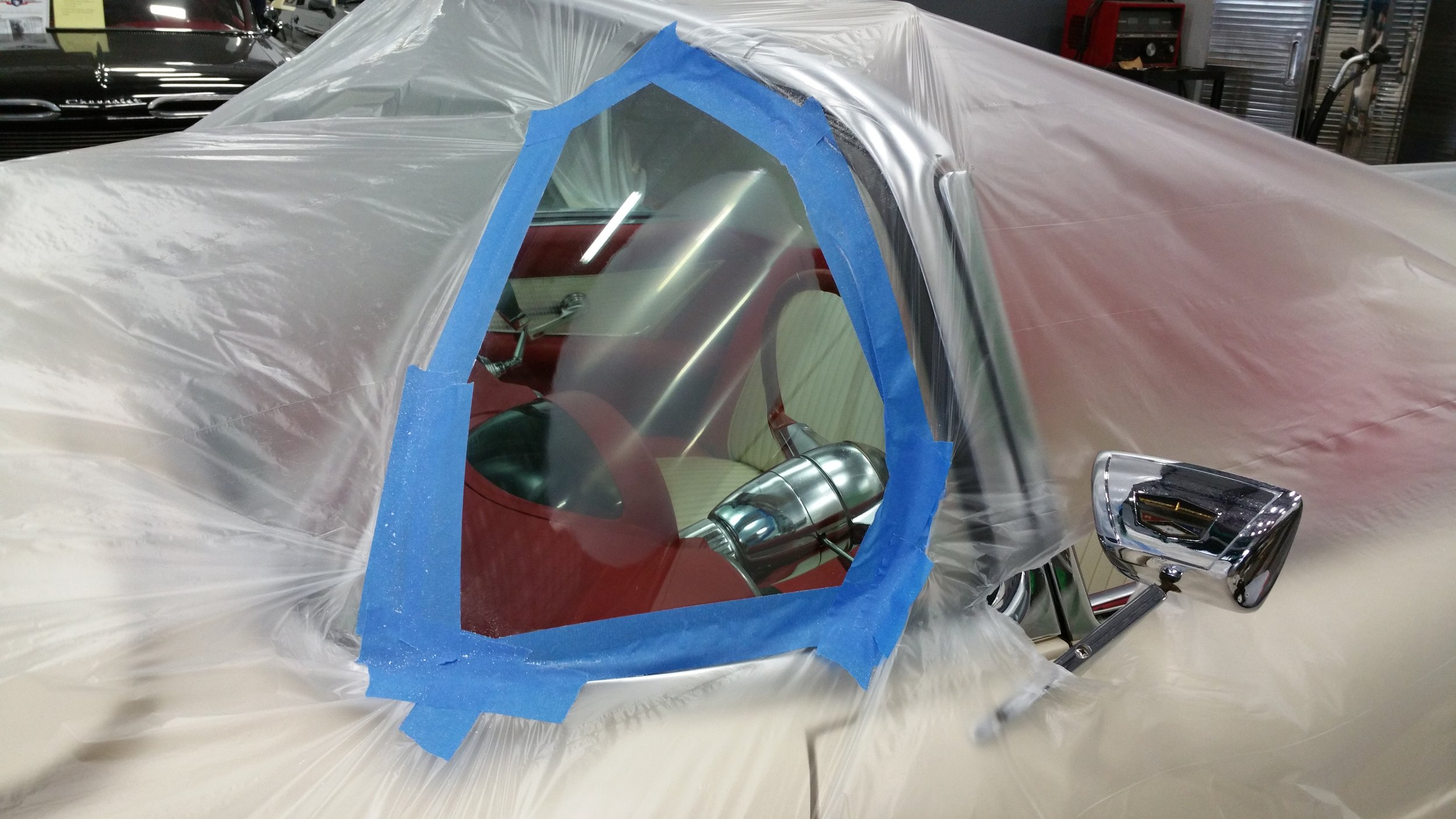 A vintage car undergoing restoration with plastic and tape covering the body. A window opening is exposed, revealing the red and beige interior. A side mirror with chrome finish is also visible.