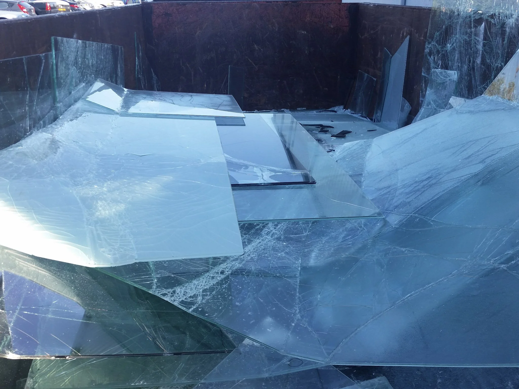 Shattered glass pieces stacked in a container, with some fragments scattered, against a background of parked cars.
