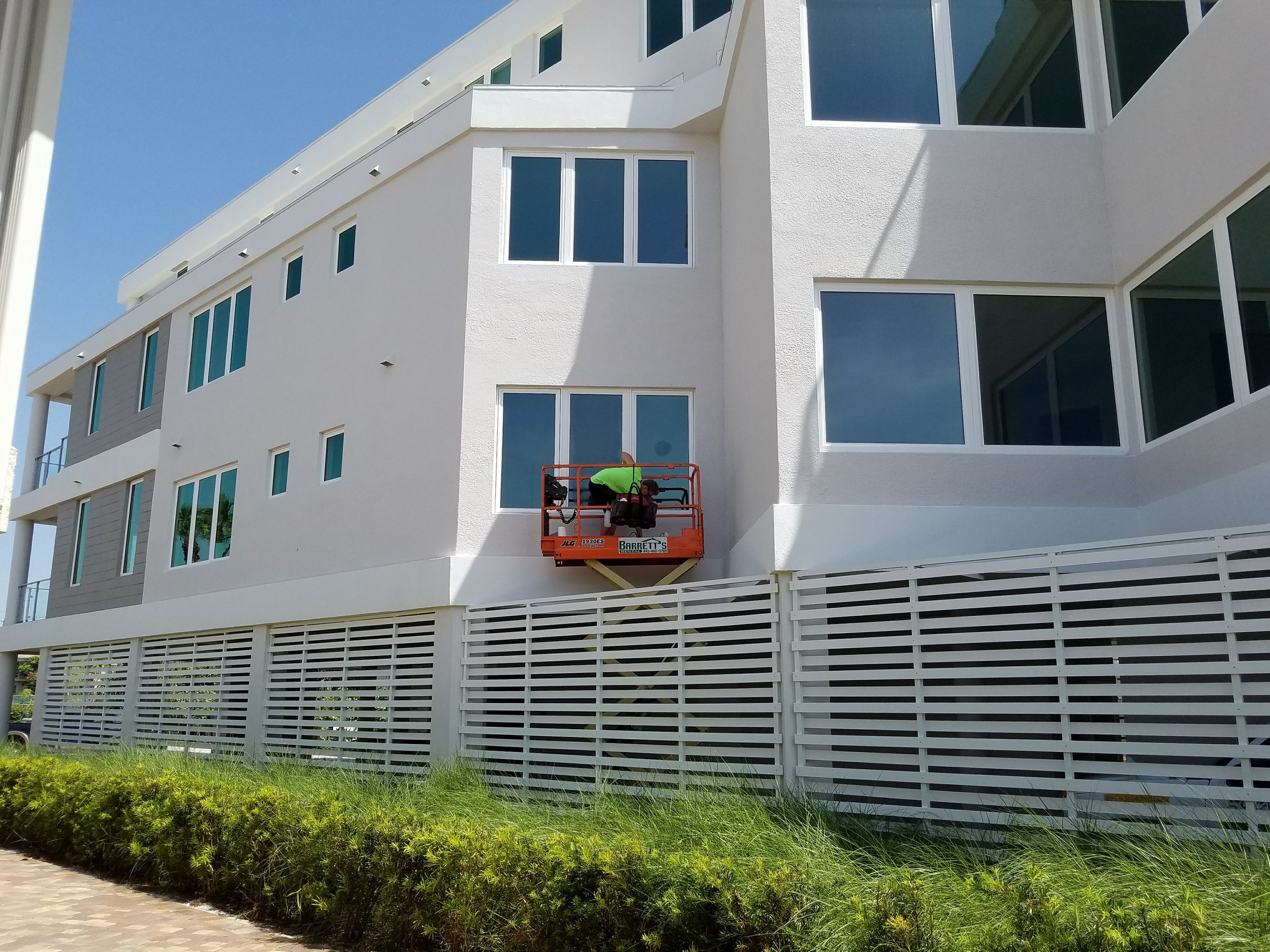 Workers on a lift inspecting windows of a modern white building with large glass panels on a sunny day.