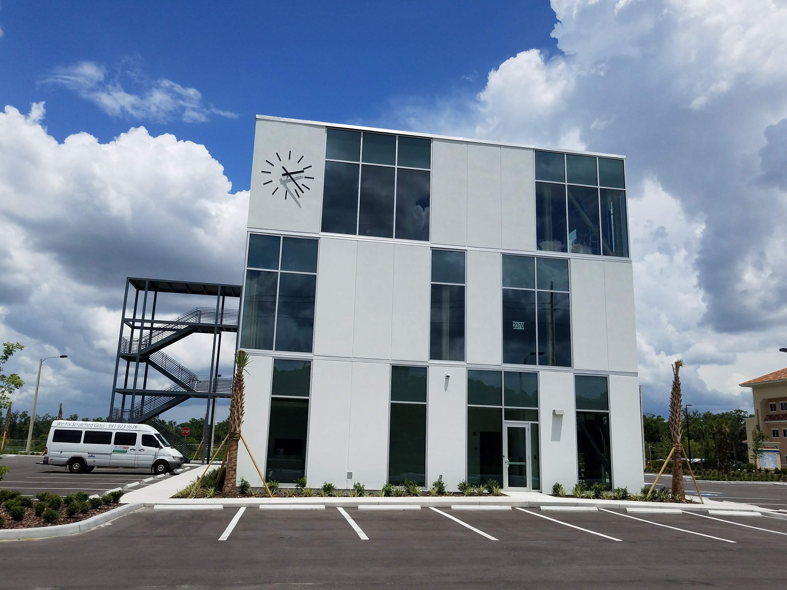 A modern three-story building with large windows and a clock face on the upper corner, located in an empty parking lot under a partly cloudy sky.