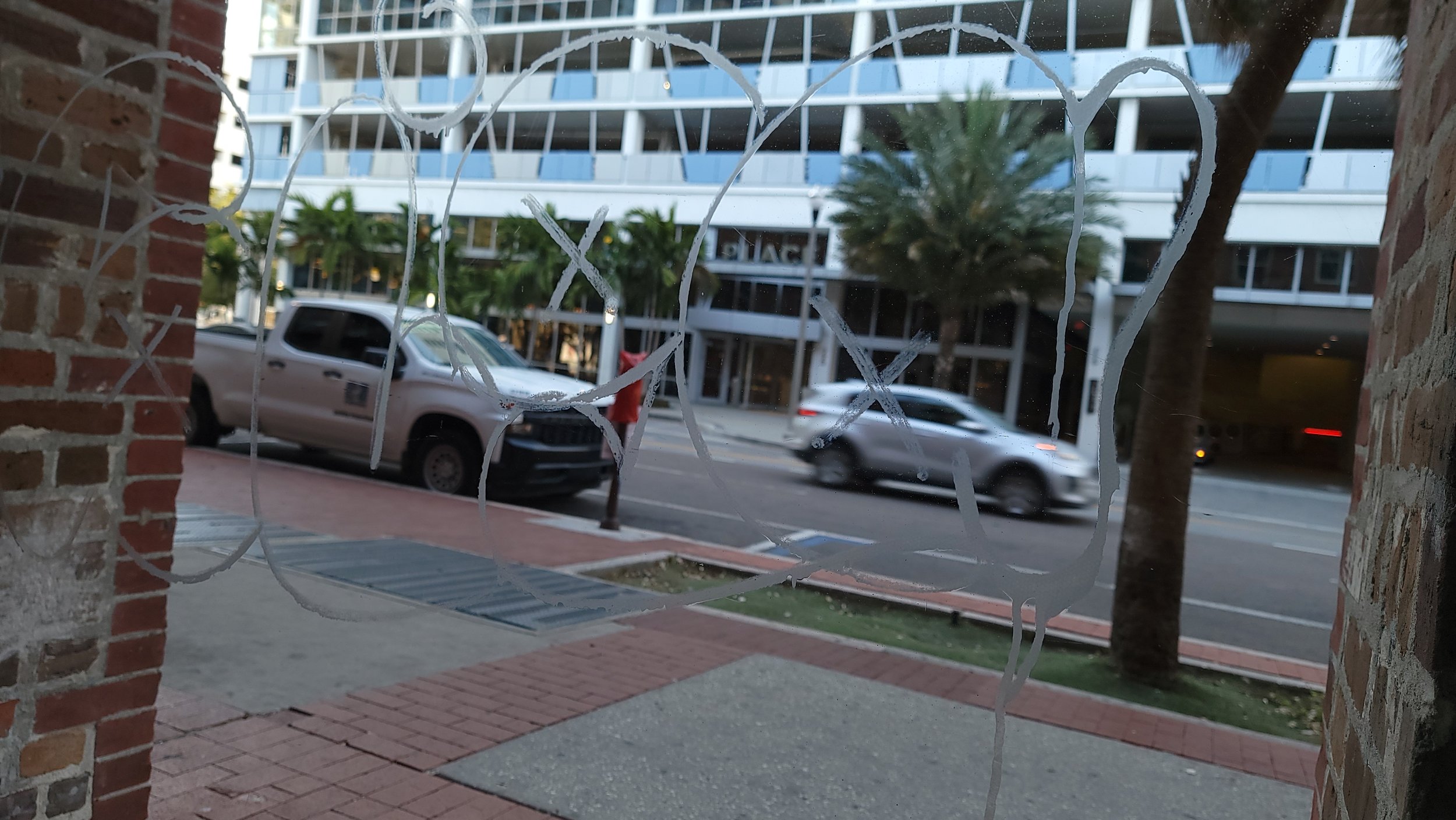 Graffiti on a glass window with a street view in the background, showing a white pickup truck and a modern building.