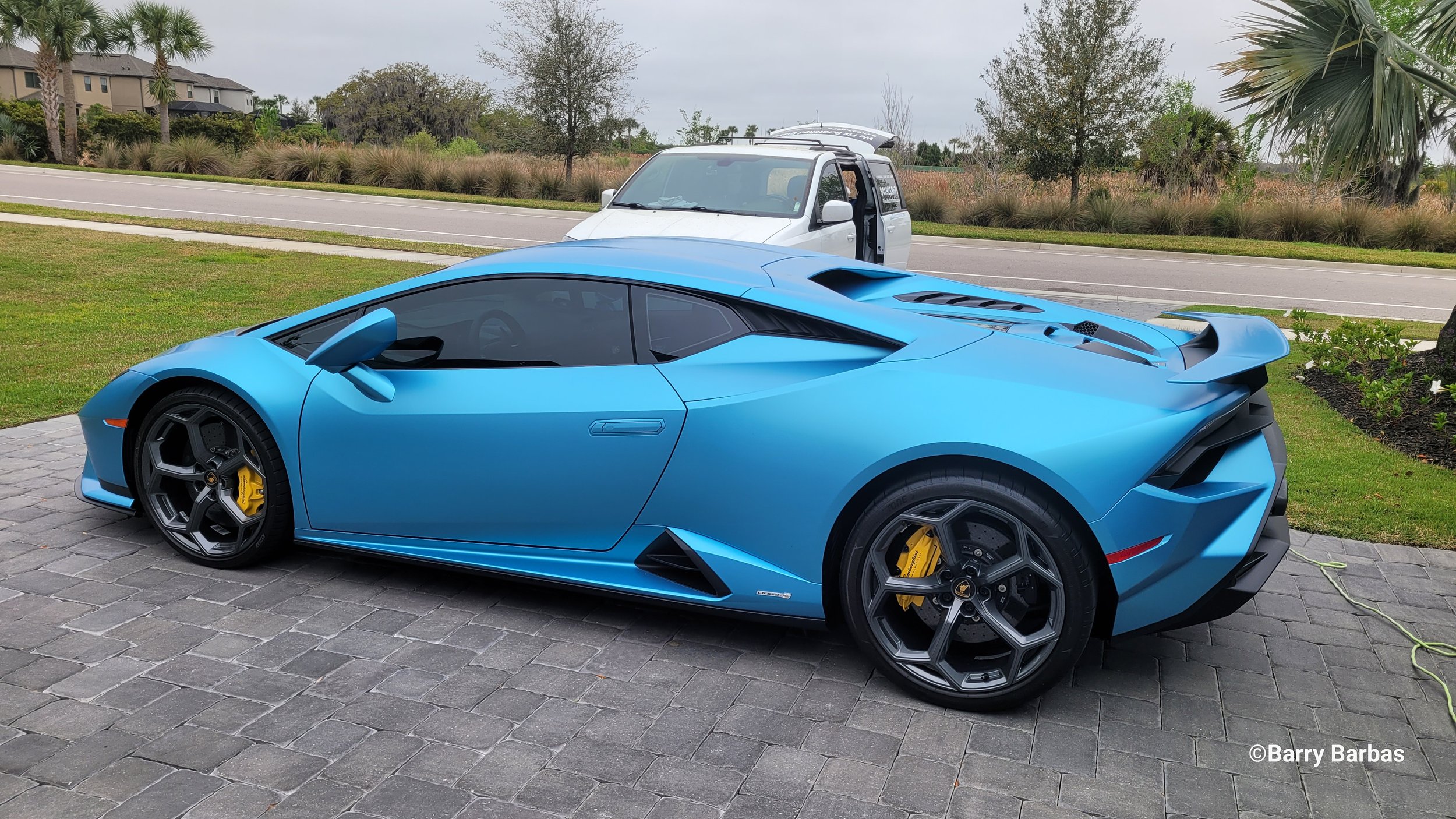 Blue sports car parked on a cobblestone driveway with a white Glass Restoration van nearby and greenery in the background.