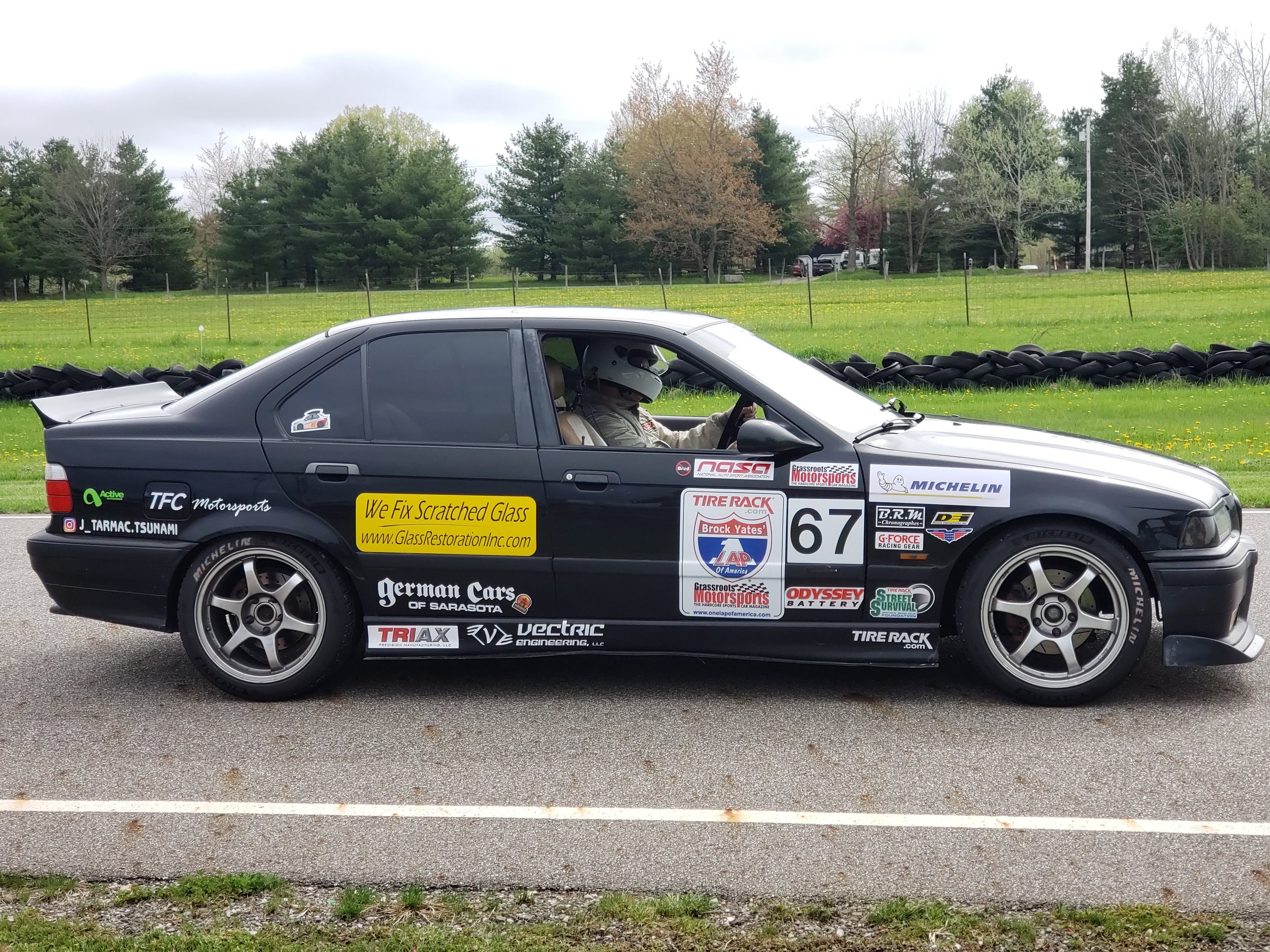 A black sports sedan with various sponsor stickers, including "We Fix Scratched Glass" and "German Cars of Sarasota," is parked on a track with a driver inside. The car is numbered 67 and features aftermarket wheels and a spoiler. Trees and a grassy