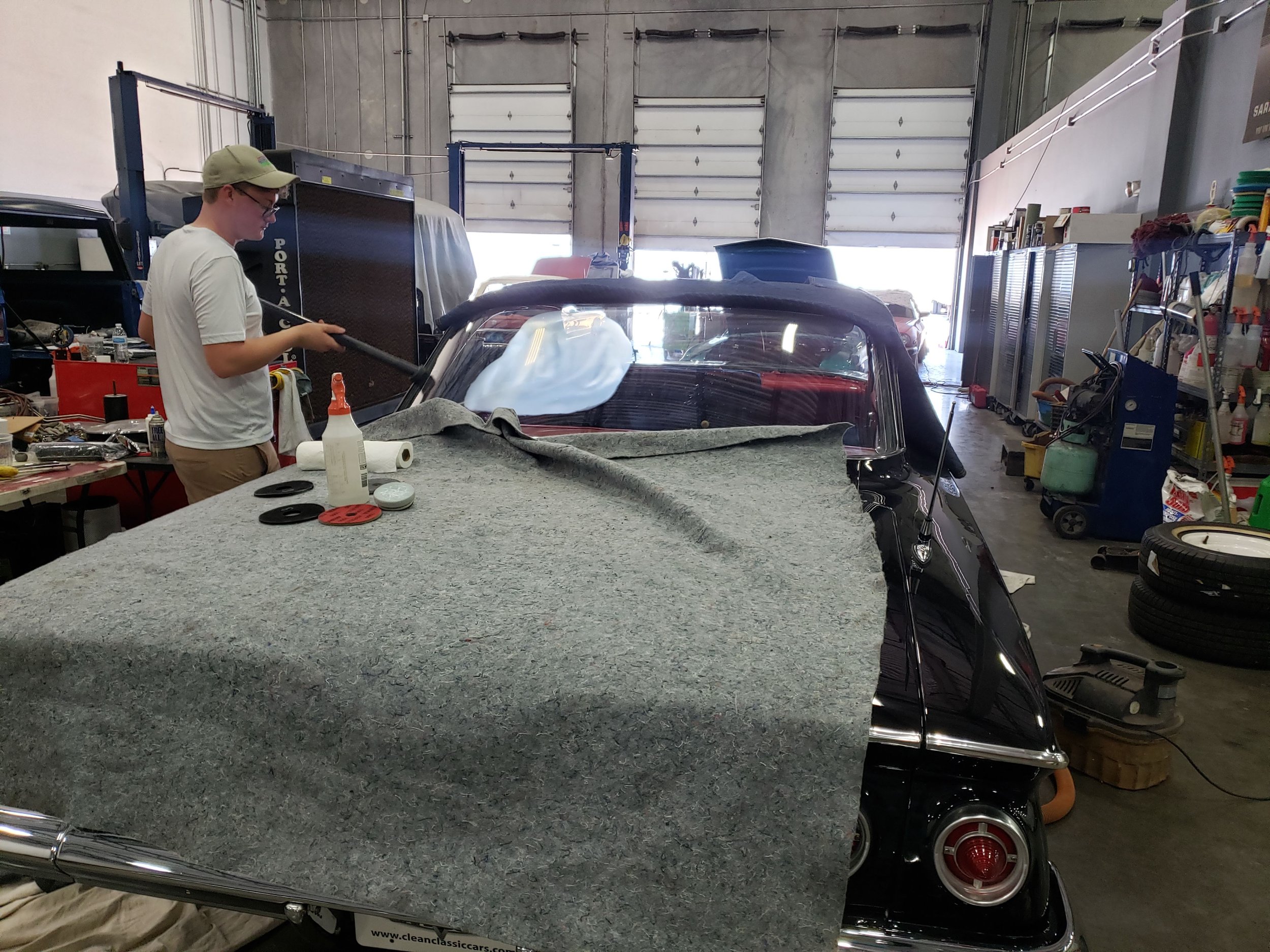 Person working on a classic car in a garage, with tools and equipment visible, and a gray cloth covering part of the car's rear.