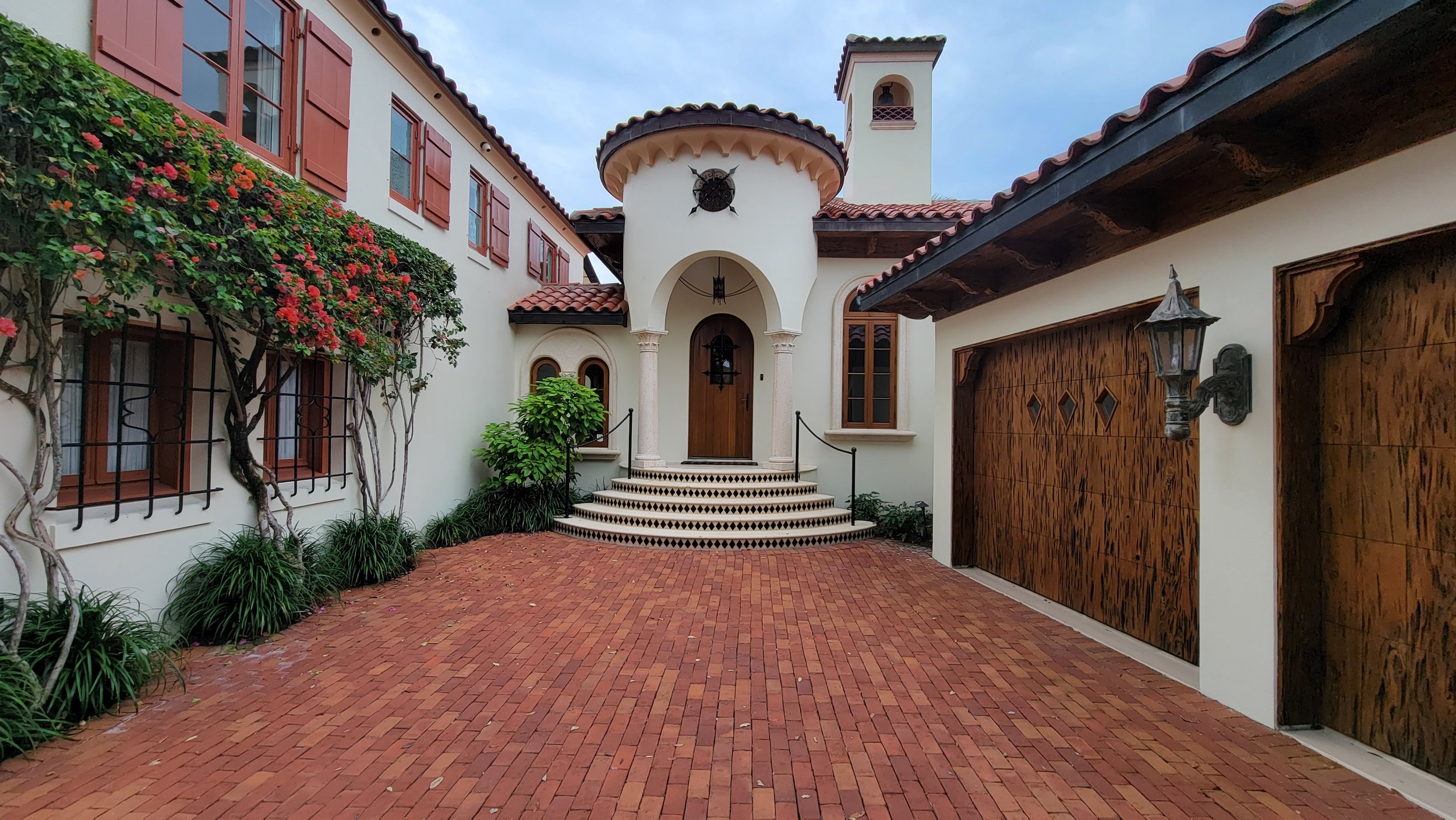 Spanish-style home with arched entryway, red tiled roof, and wooden garage doors. Brick courtyard with flowering vines on the facade.