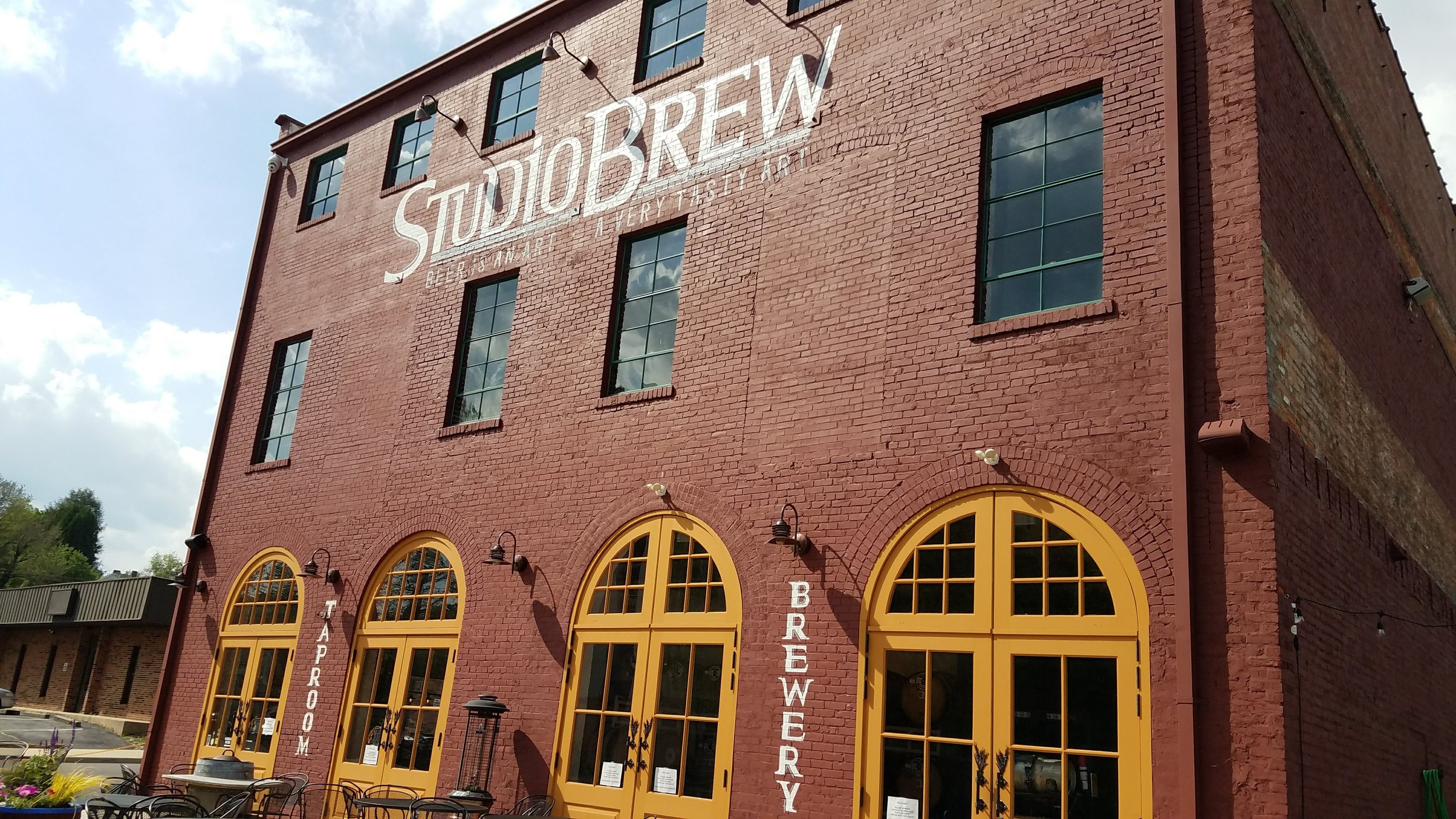 Exterior of a red brick building labeled "Studio Brew," featuring large arched yellow doors and windows, with outdoor seating visible.