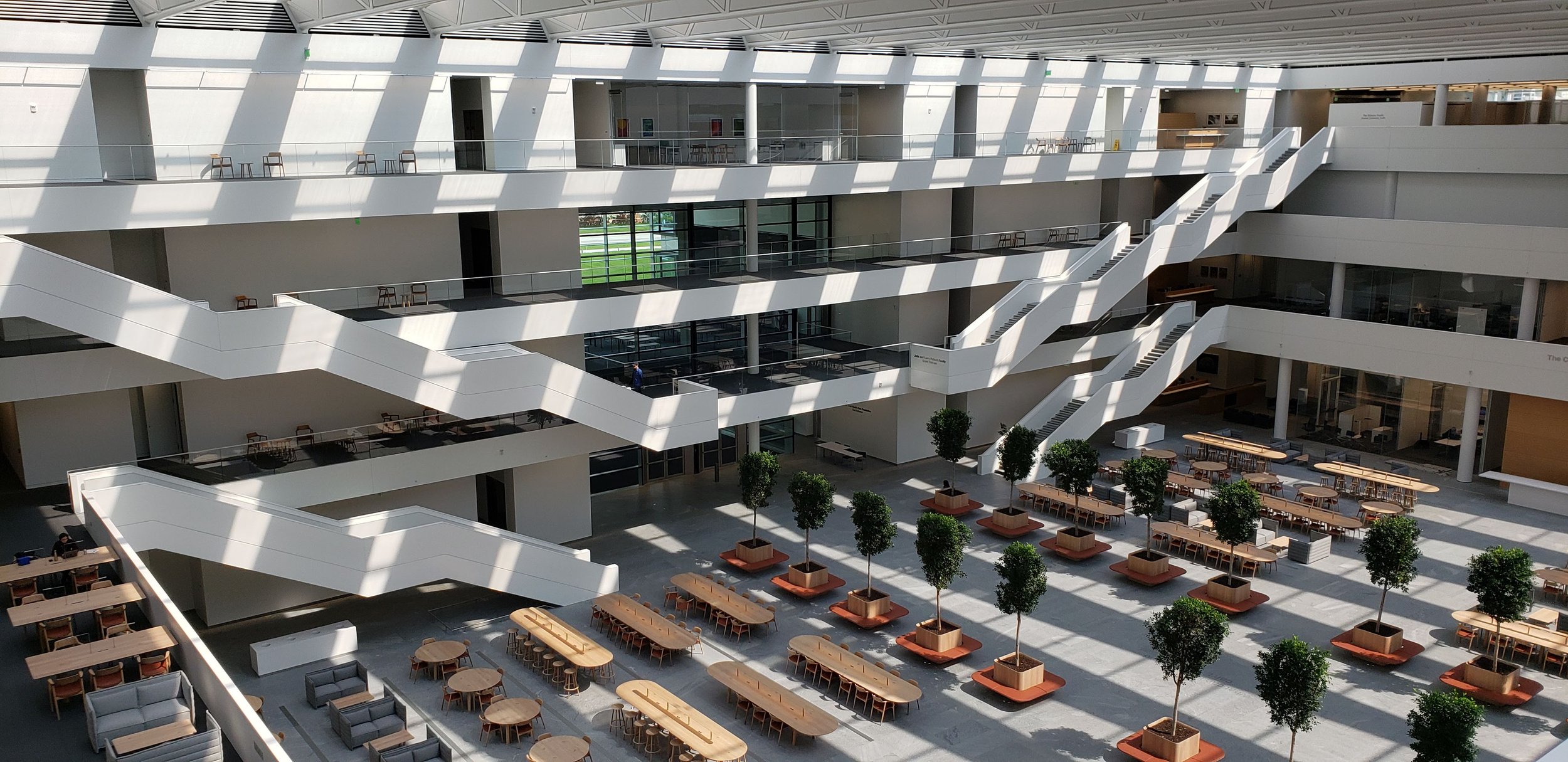 Spacious, modern interior with multiple levels and zigzagging staircases, featuring high ceilings, ample natural light, tables, chairs, and indoor potted trees.