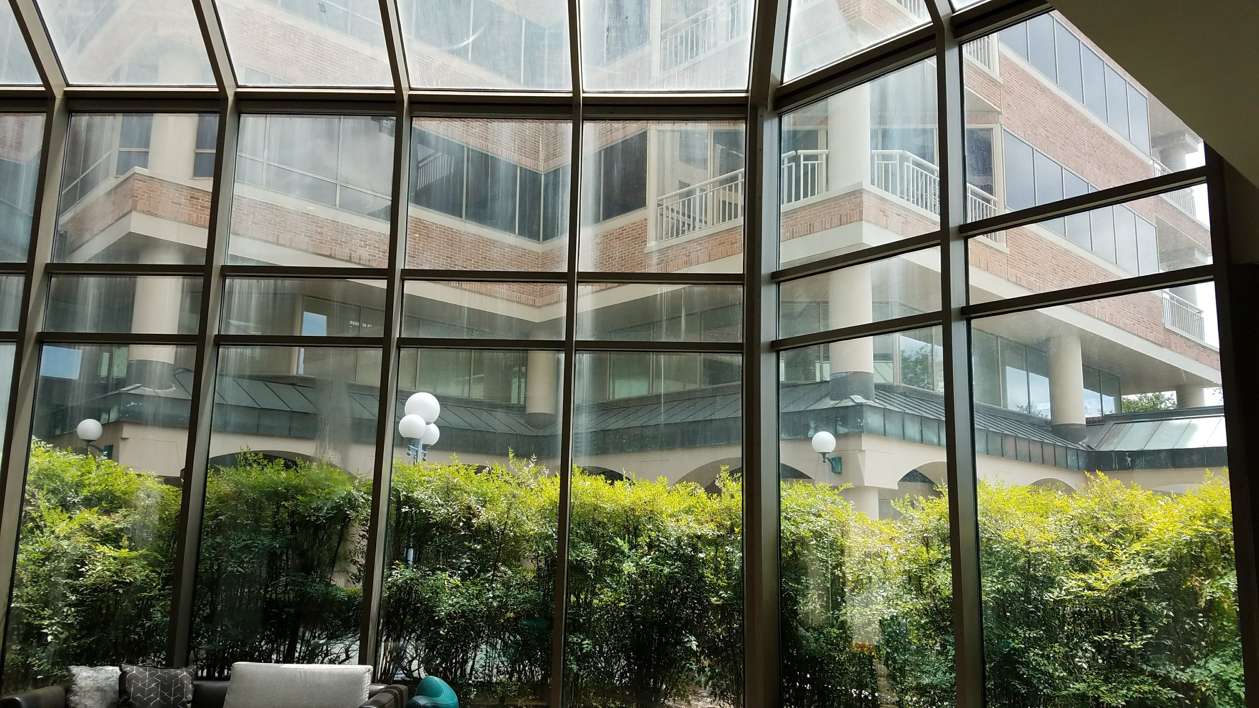 Interior view through large glass windows with a brick building, greenery, and outdoor lamps in the background.