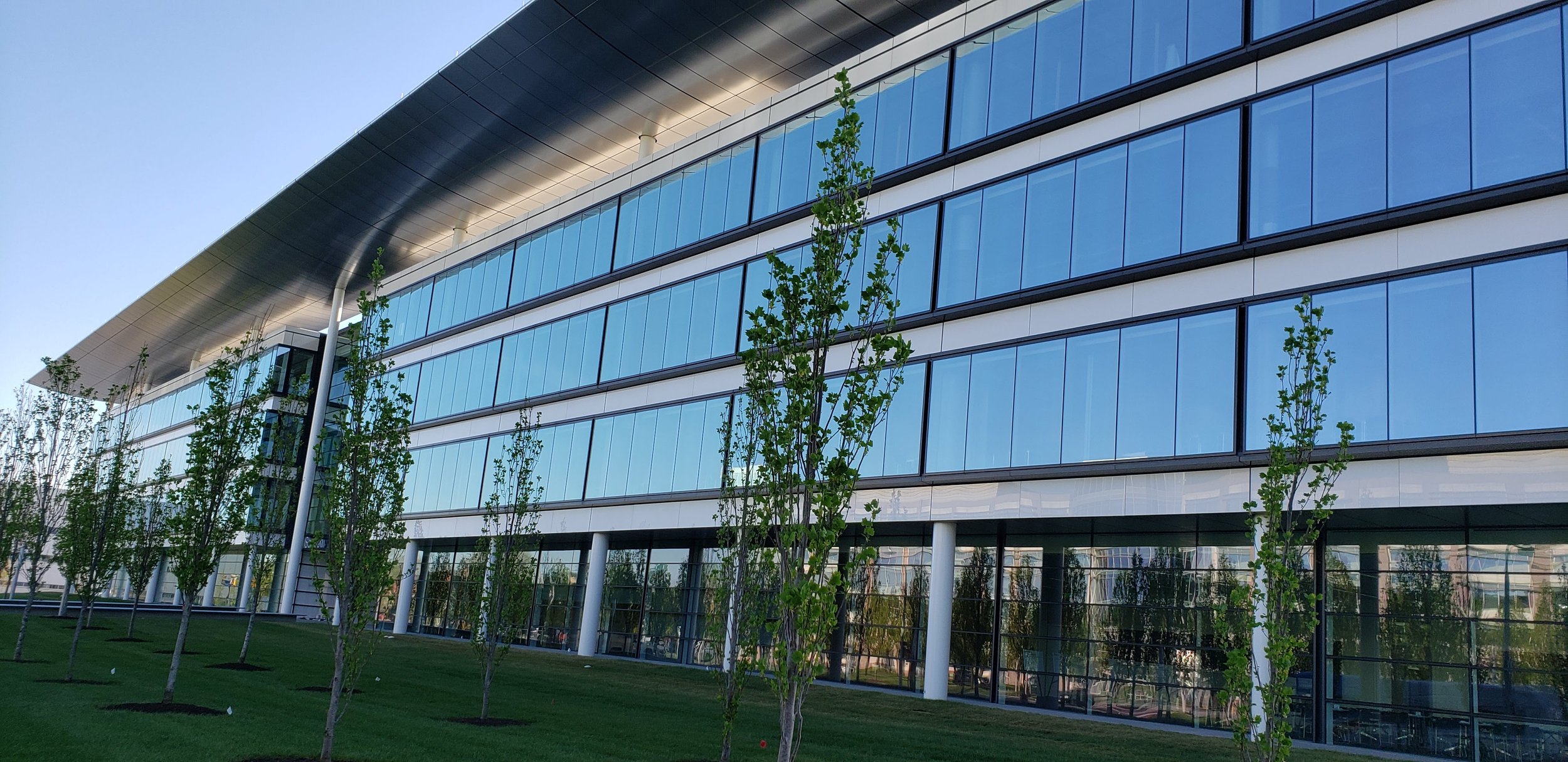 Modern office building with large glass windows and a rooftop overhang, surrounded by young trees and green lawn.