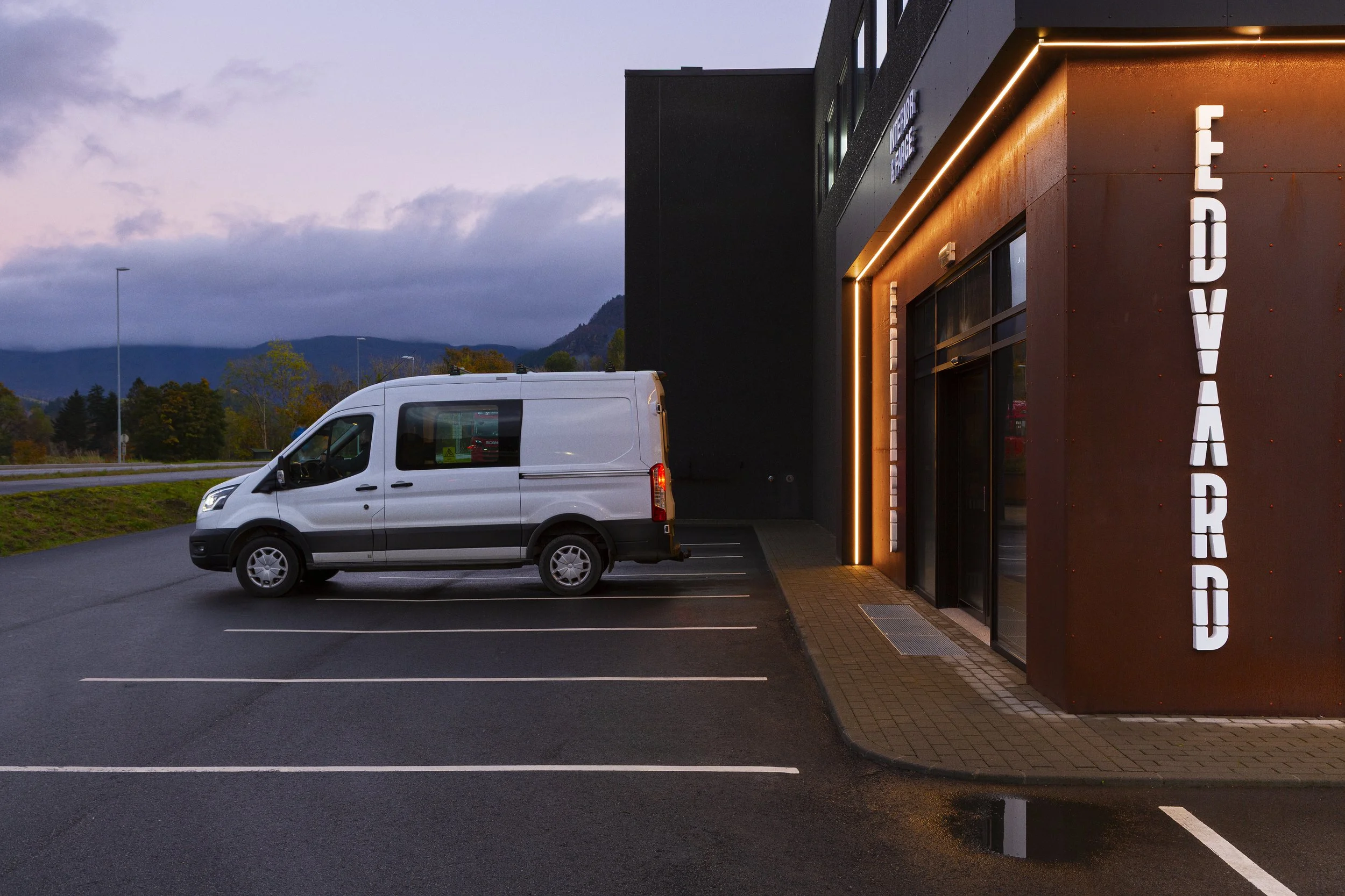 Foto av en tom parkeringsplass foran Ljoshallbygget med en stor vertikal skilt som sier 'EDVARD', og en hvit varebil parkert nær inngangen, i skumringstid med overskyet himmel.