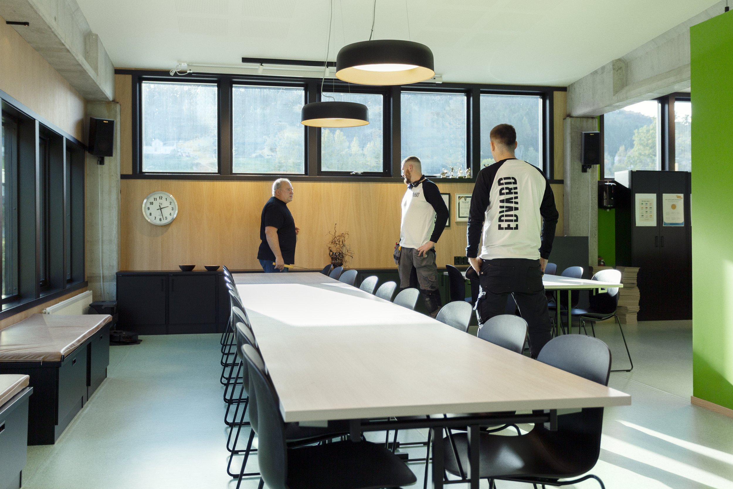 Tre personer i et konferanserom, to av dem med «EVOARD» på jakker, en annen person står ved et bord. Rommet har store vinduer og moderne belysning.