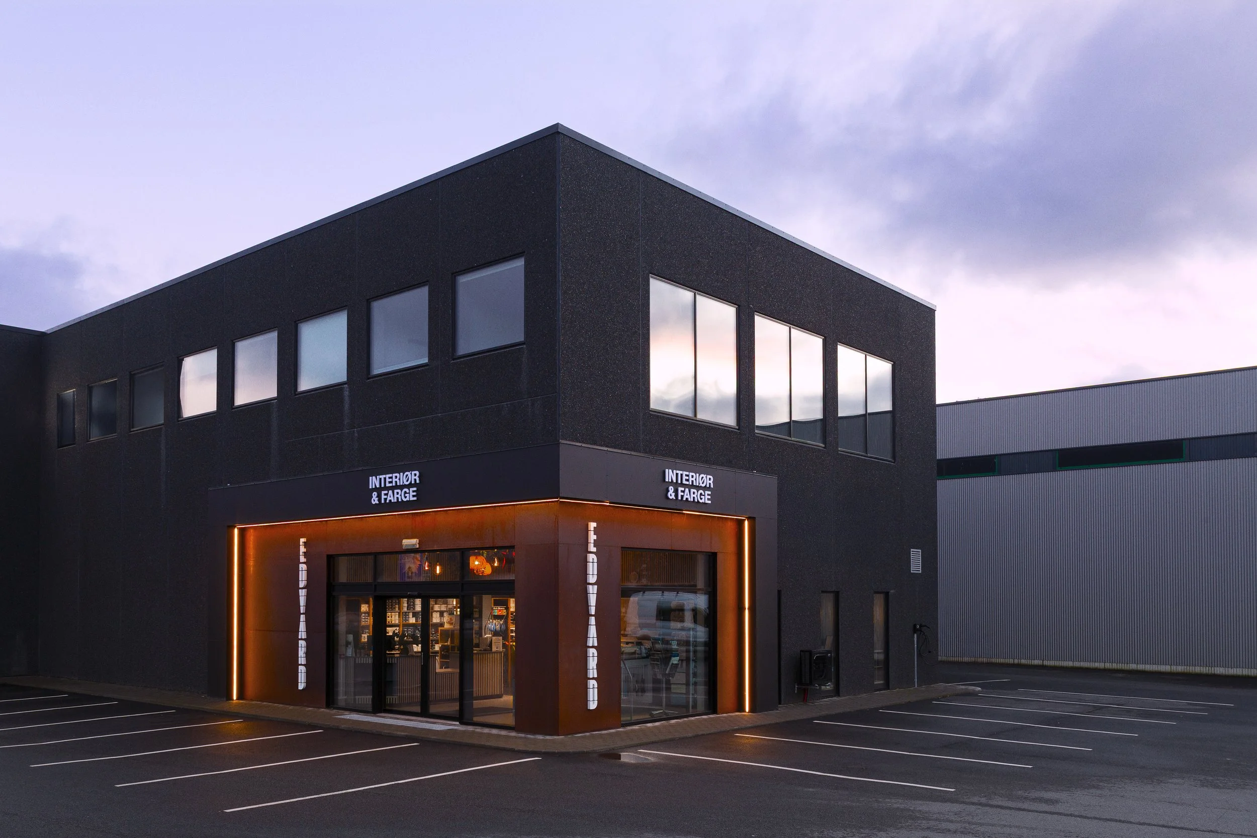 En moderne toetasjes butikkbygning med skilt som sier 'Interior & Farge' og 'Lieverd', med parkeringsplass foran. Bygningen har store vinduer og er opplyst i vinduene. Dette er tatt om kvelden eller tidlig morgen.