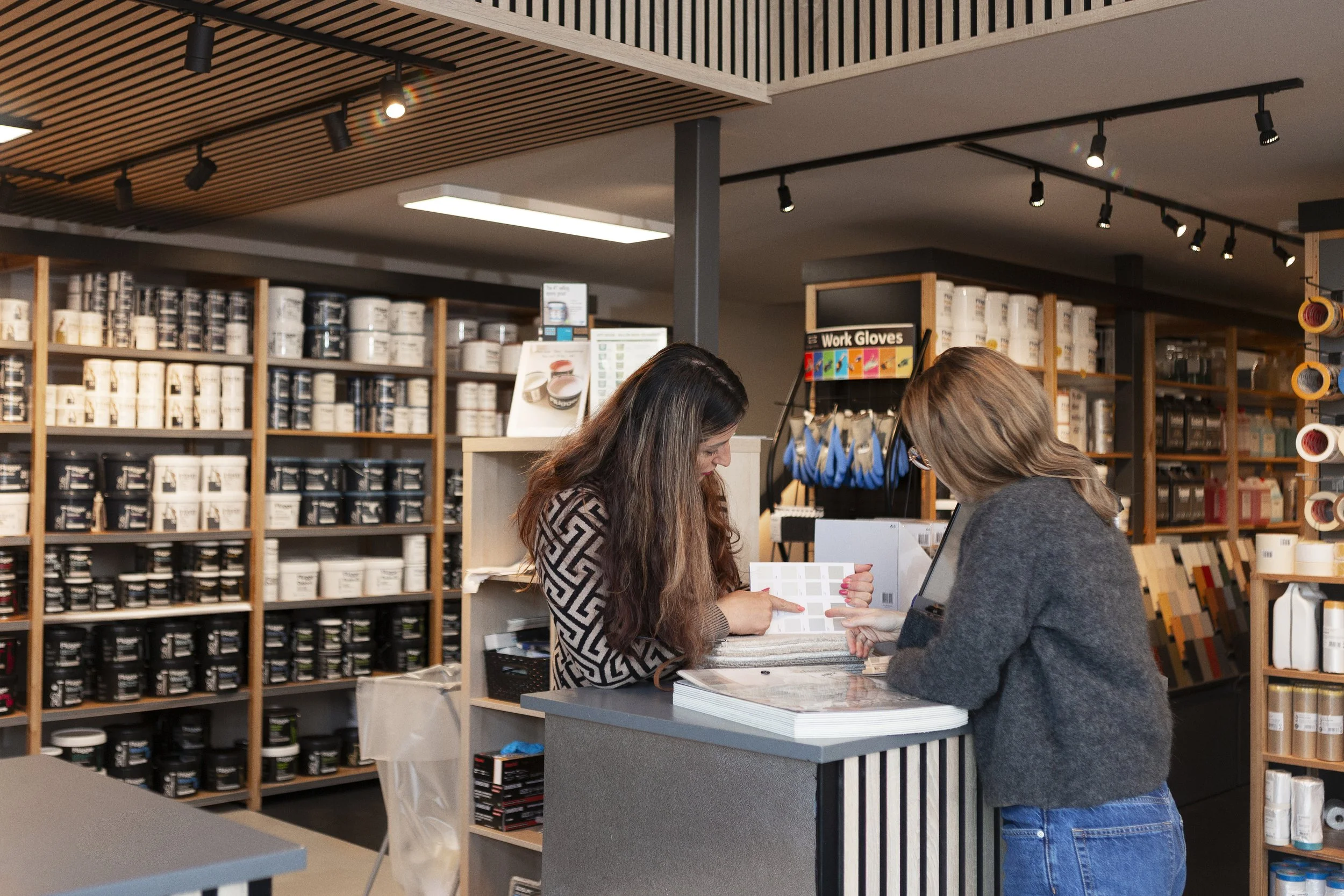 To kvinner står ved en butikkdisk, ser på fargeprøver i et butikkmiljø fylt med hyller av malingbokser og byggemateriell.