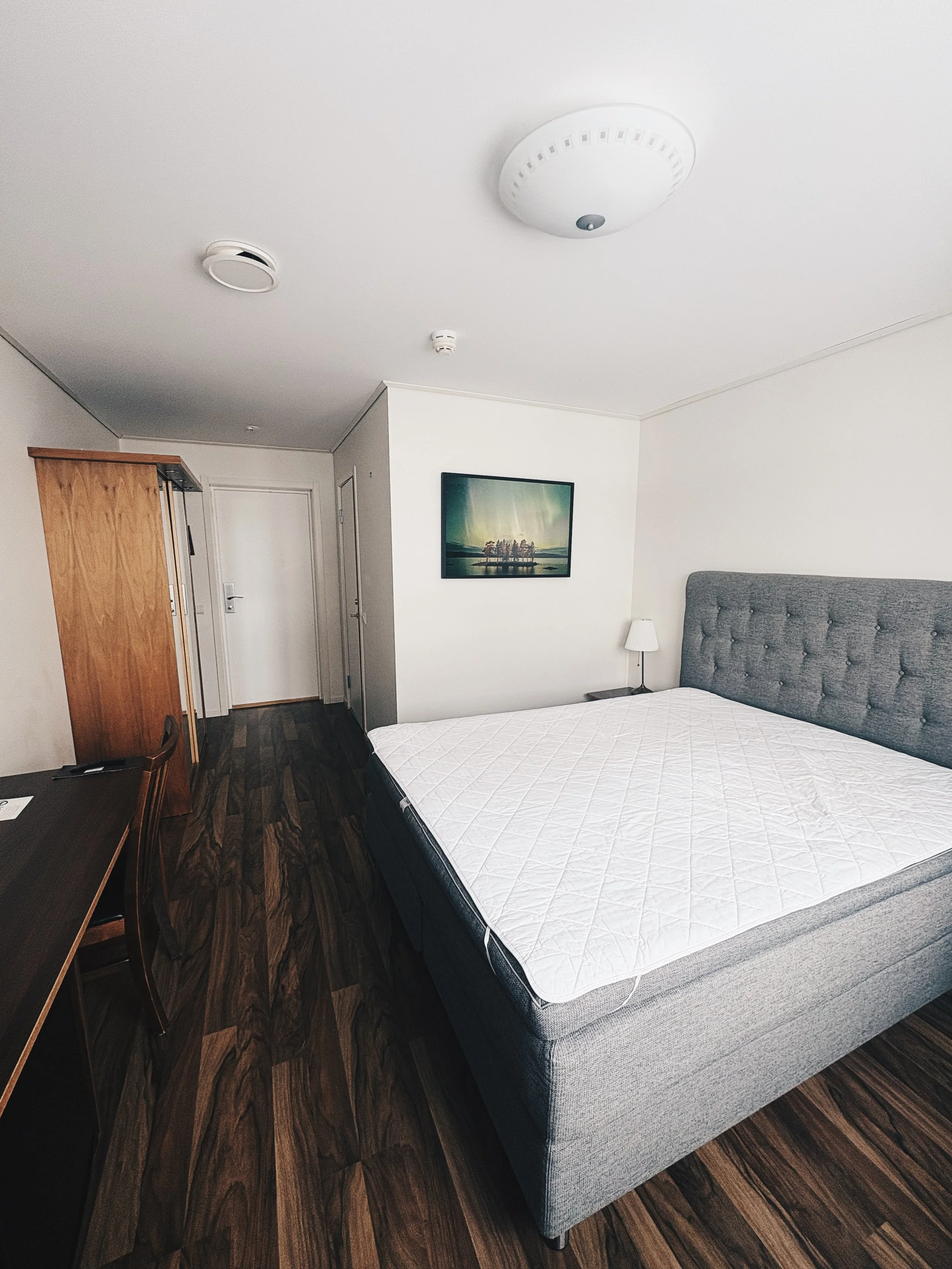 Hotel room with a grey upholstered bed, white mattress, wooden floor, a desk with a chair, a wardrobe, a wall-mounted television, a small nightstand with a lamp, and a door.