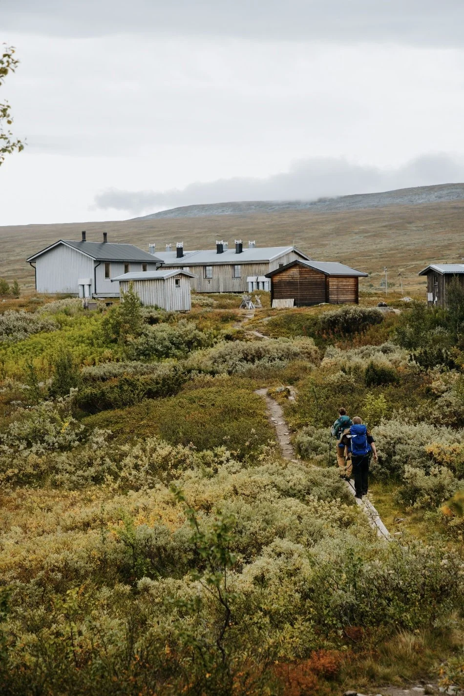 Two hikers with backpacks walking along a narrow path through green and brown shrubbery toward a small cluster of rustic wooden houses, with rolling hills and cloudy sky in the background.