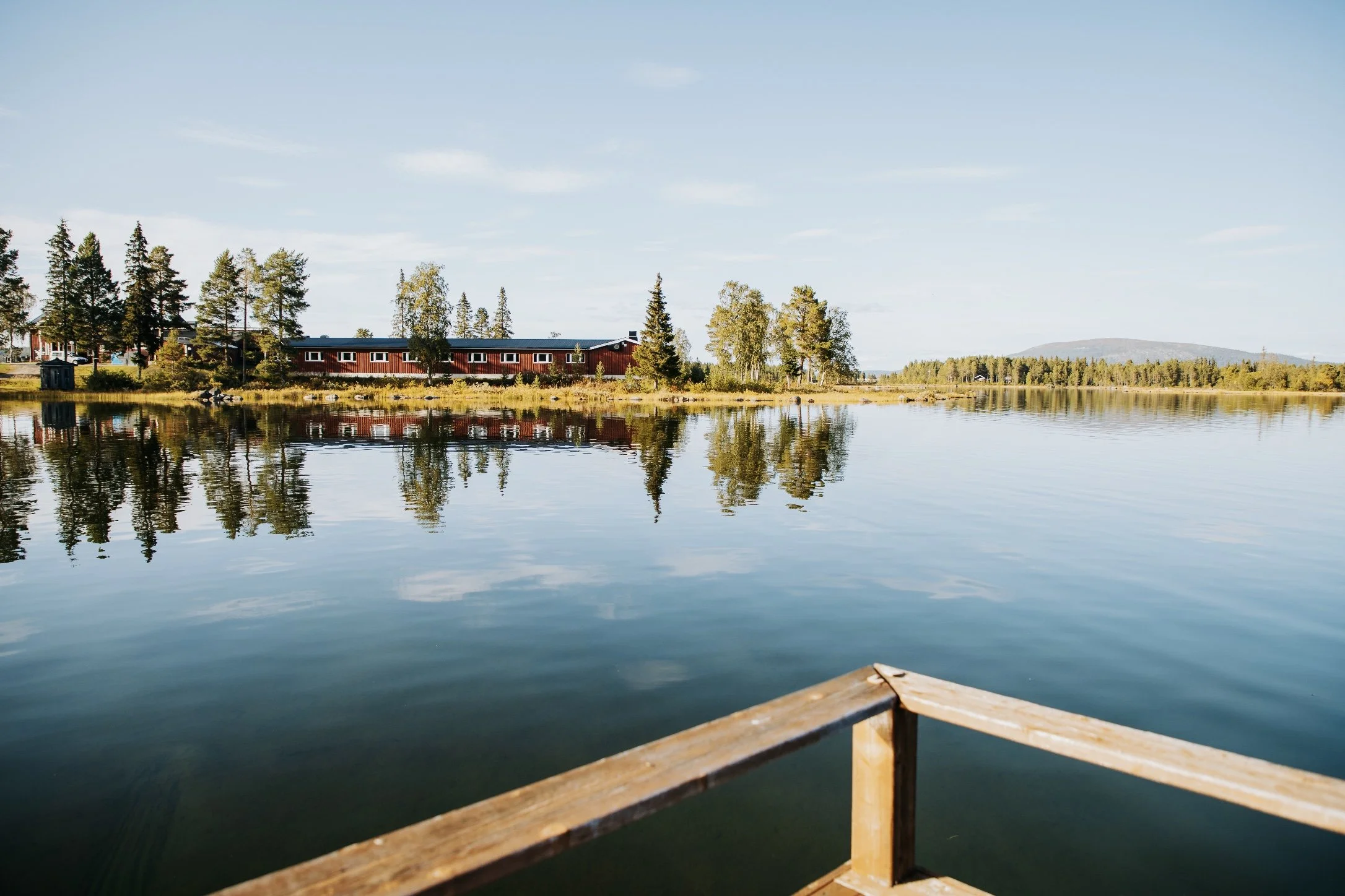 View of a calm lake with trees and houses on the shoreline, blue sky with light clouds.