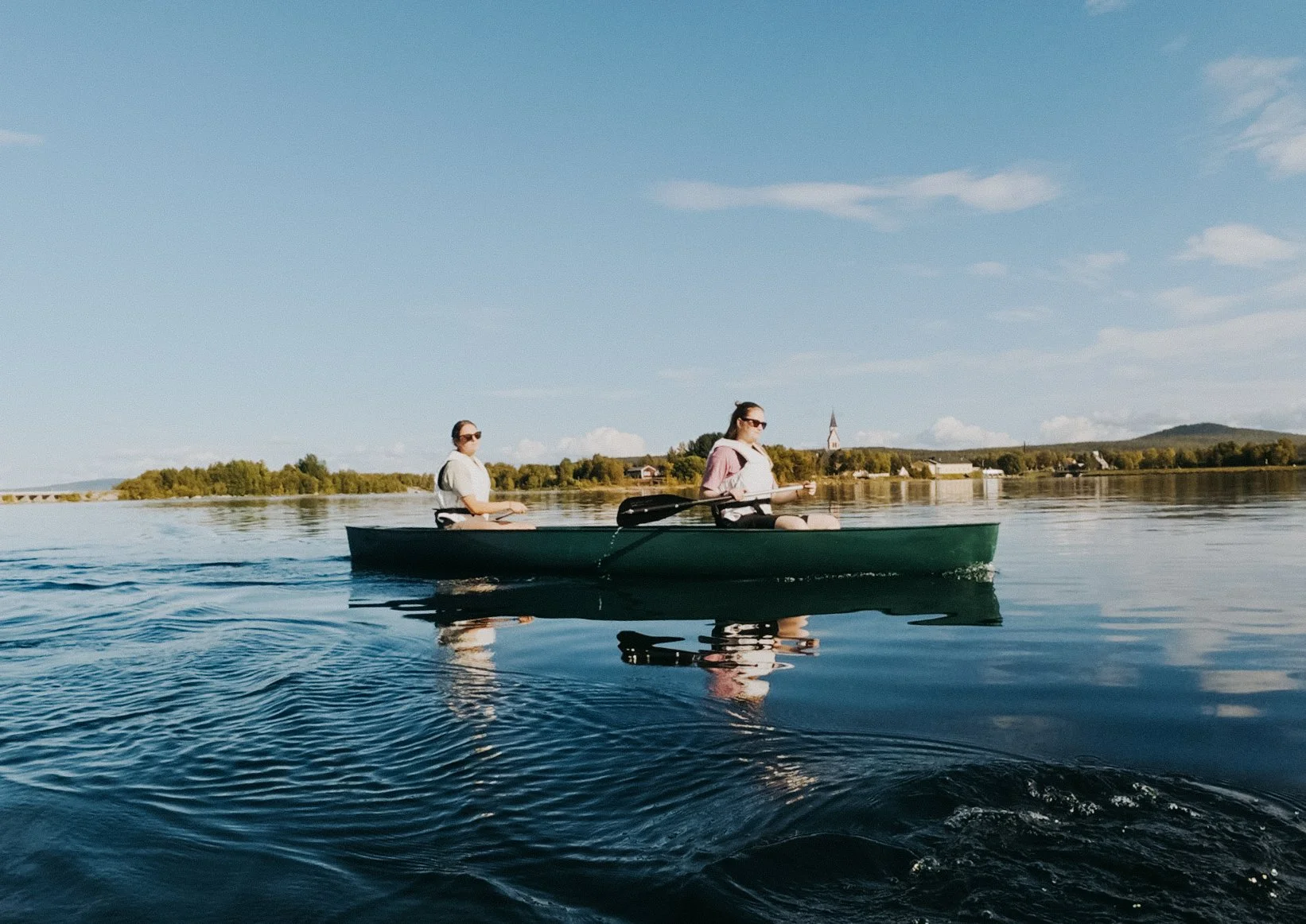 Two women in a green canoe paddling on a calm lake under a blue sky with a few clouds, with a village and hills in the background.