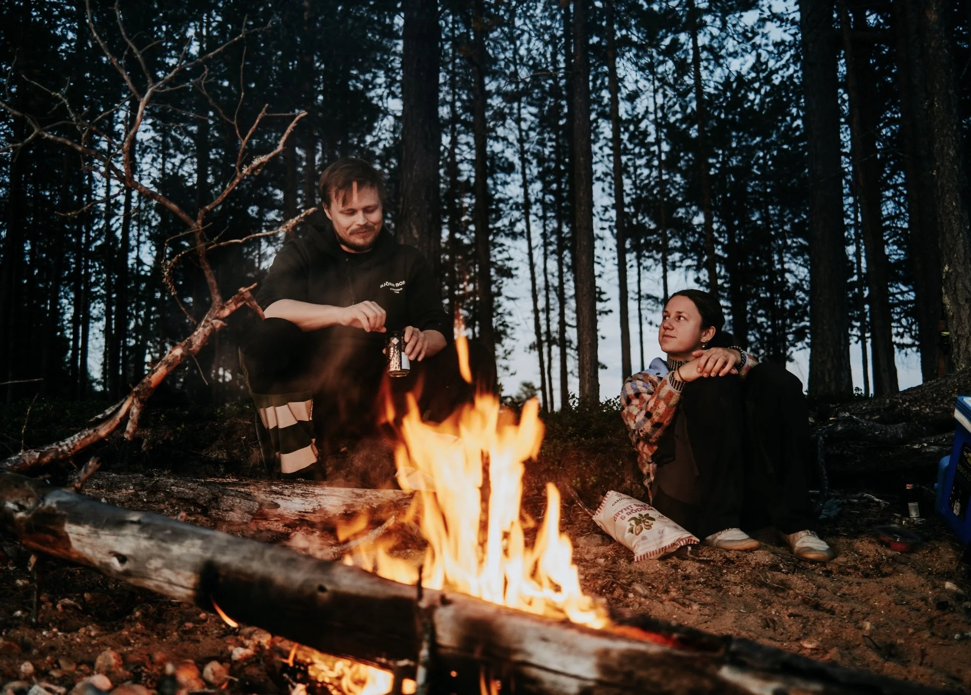 A man and woman sitting around a campfire in a forest clearing at dusk.