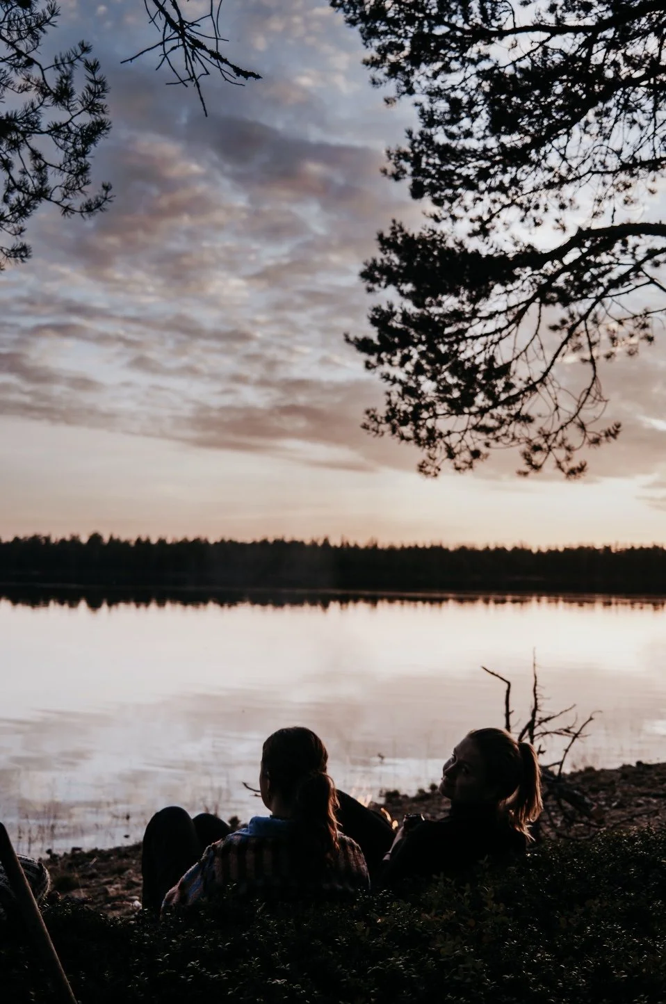 Two women sitting on the ground near a lake at sunset, with trees framing the scene and a cloudy sky overhead.