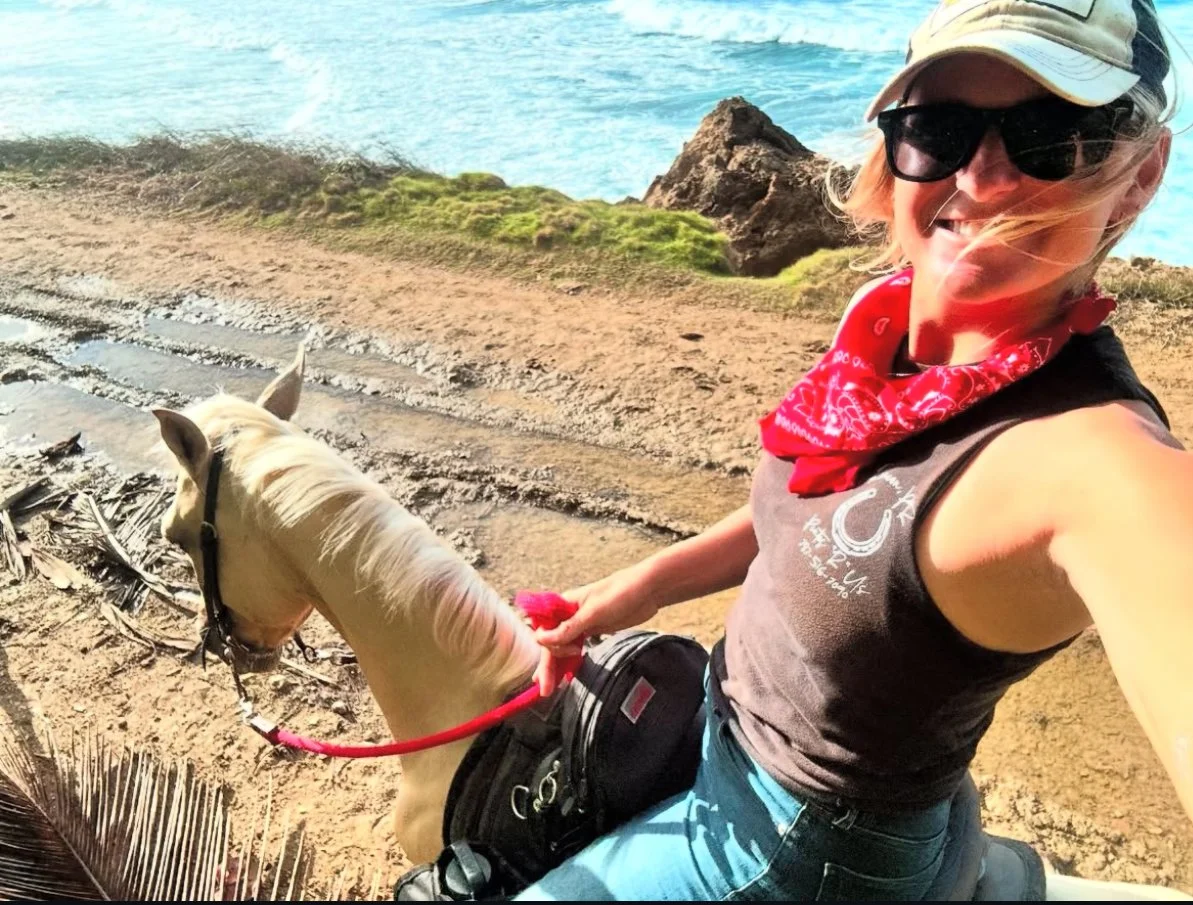 A woman with sunglasses, a cap, and a red bandana around her neck riding a cream-colored horse along a sandy beach with rocks and the ocean in the background.