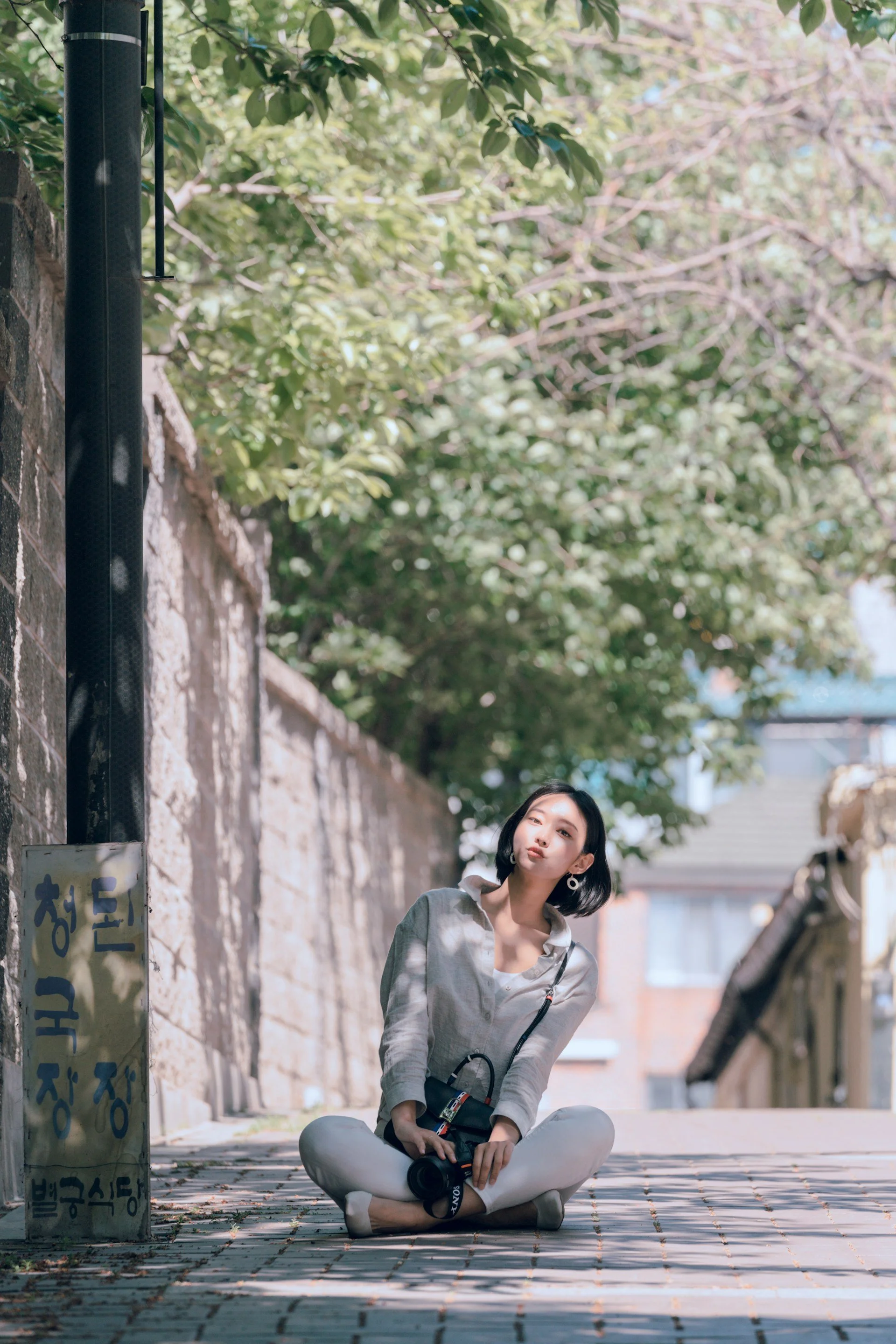 A woman sitting cross-legged on a cobblestone street, holding a camera, with a stone wall and leafy green trees in the background.