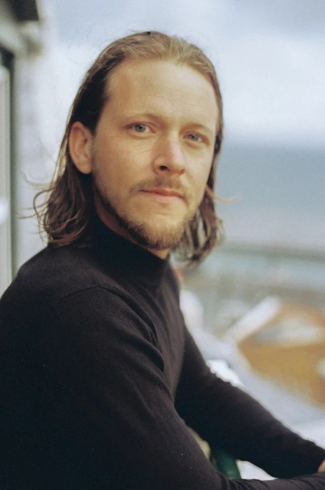 A man with shoulder-length brown hair and a beard, wearing a black shirt, is looking at the camera with a slight smile. The background shows a blurred outdoor scene with a cloudy sky and water.