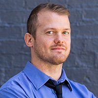 A man with short brown hair and a beard, wearing a blue shirt and a black tie, standing in front of a dark brick wall.