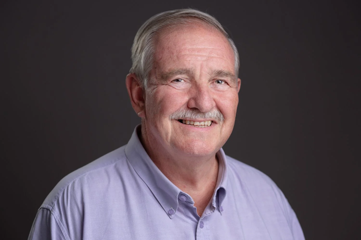 A smiling older man with gray hair and a mustache wearing a light purple button-up shirt, standing against a dark gray background.