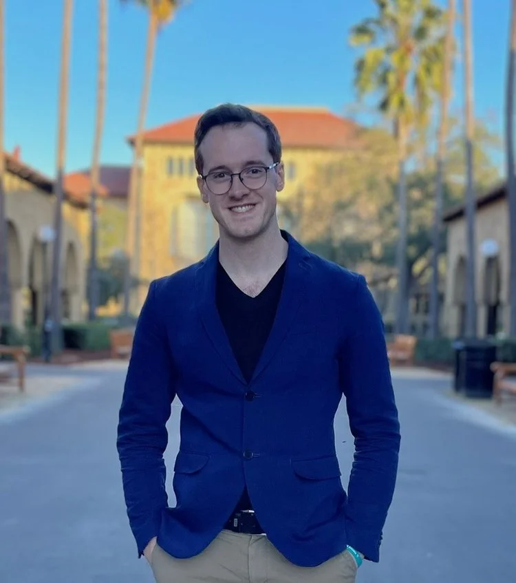 A young man with glasses, smiling, wearing a navy blazer and black shirt, standing outdoors on a sunny day with palm trees and a building in the background.